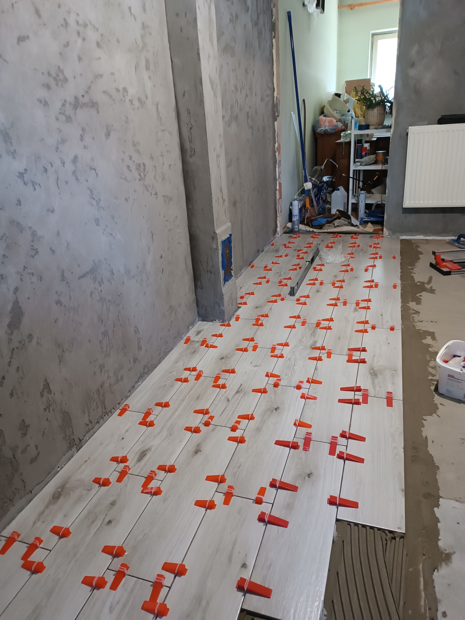 Układanie jasnych płytek podłogowych z użyciem pomarańczowych klipsów poziomujących. Widoczna surowa ściana i narzędzia glazurnicze w tle. Etap prac wykończeniowych.