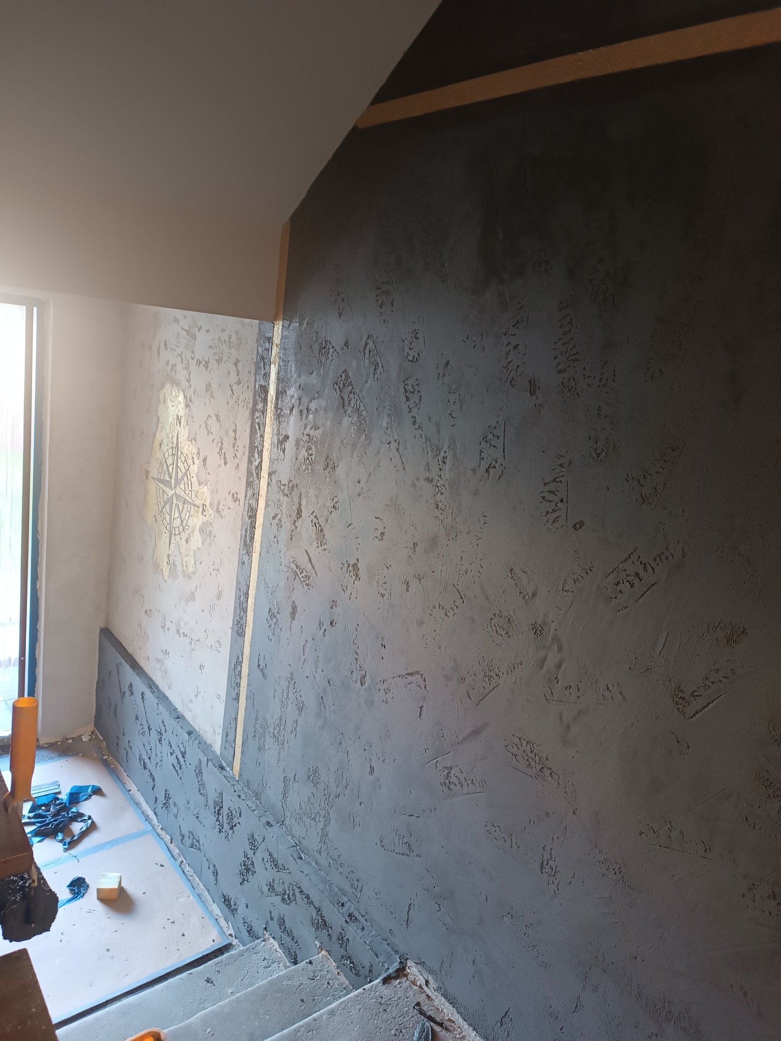 Teksturowana ściana w odcieniach szarości z motywem kompasu, oddzielona drewnianą listwą. Widoczne schody i narzędzia malarskie.