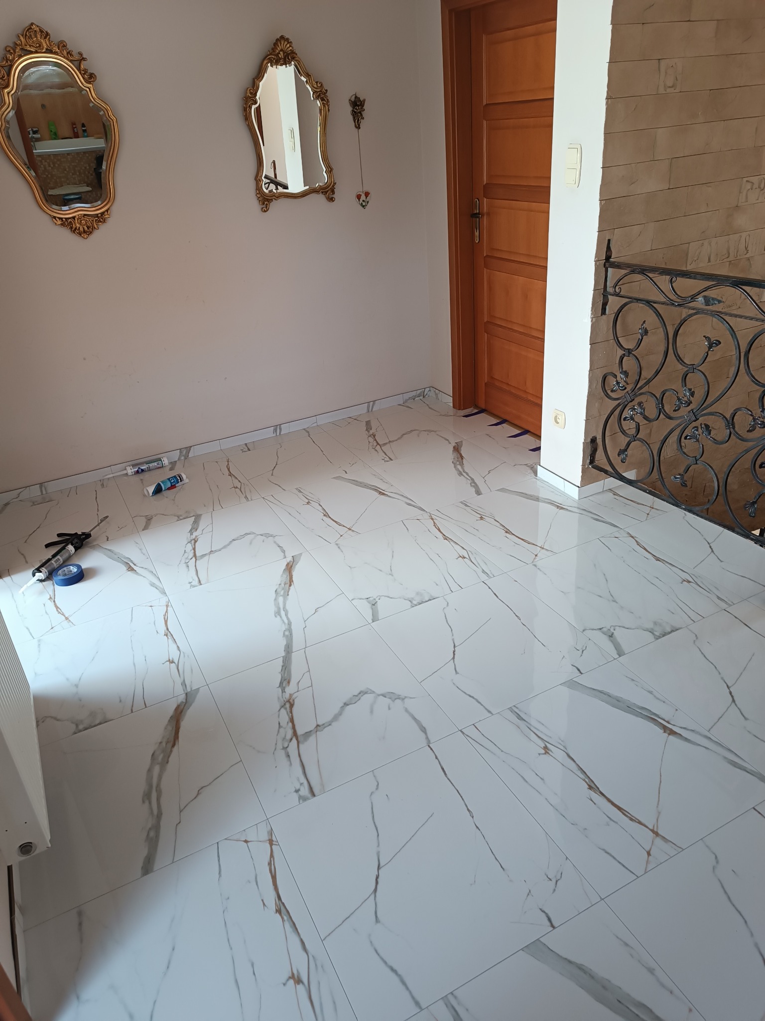 Białe płytki podłogowe z marmurowym wzorem w pokoju, widoczne narzędzia glazurnicze i dekoracyjne lustra na ścianie. Wykończone wnętrze z elementami dekoracyjnymi.