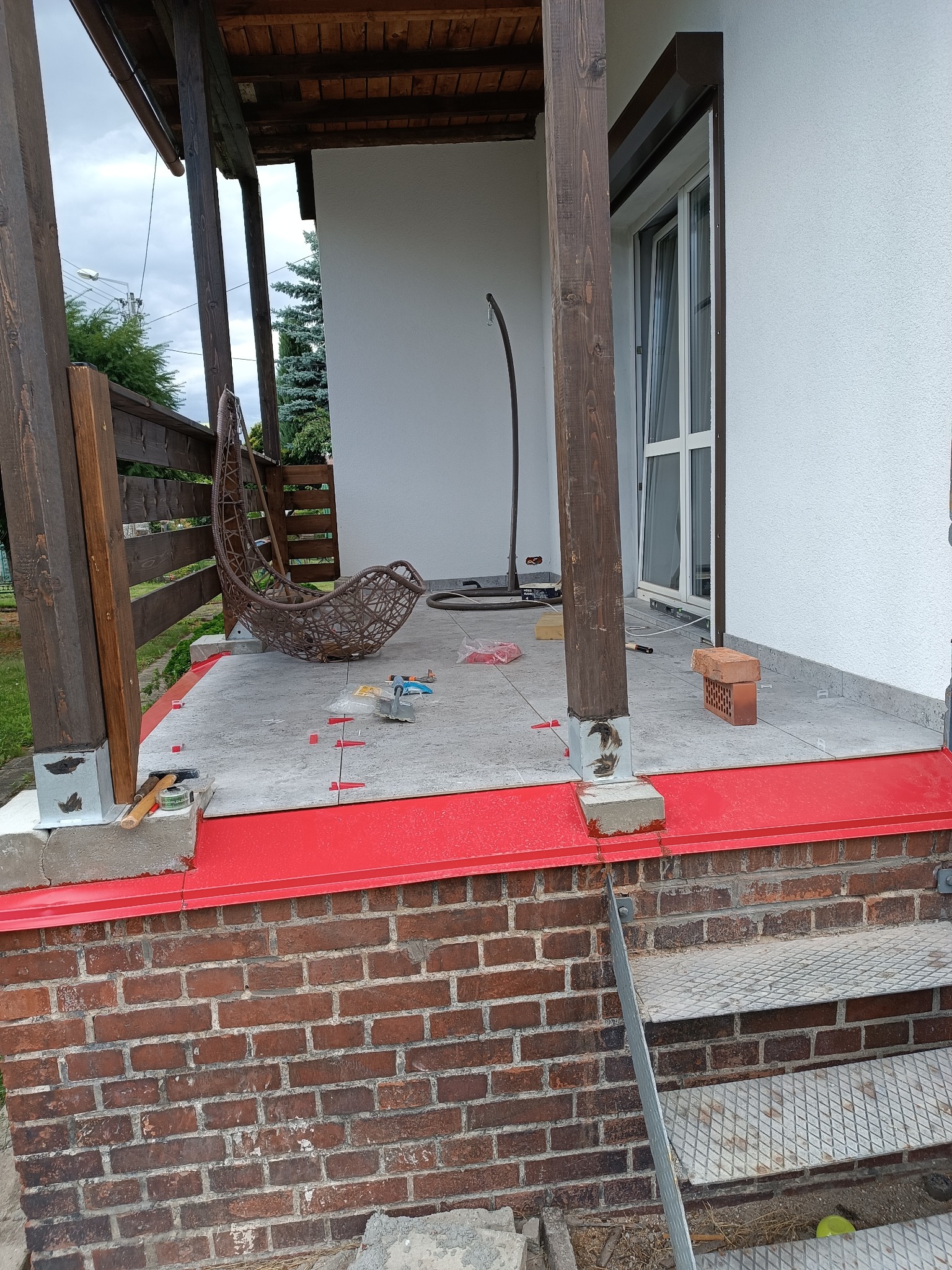 Kładzenie szarych płytek na tarasie domu z brązowymi słupami. Widoczne narzędzia glazurnicze i czerwone elementy wykończeniowe. W tle wiszące krzesło.