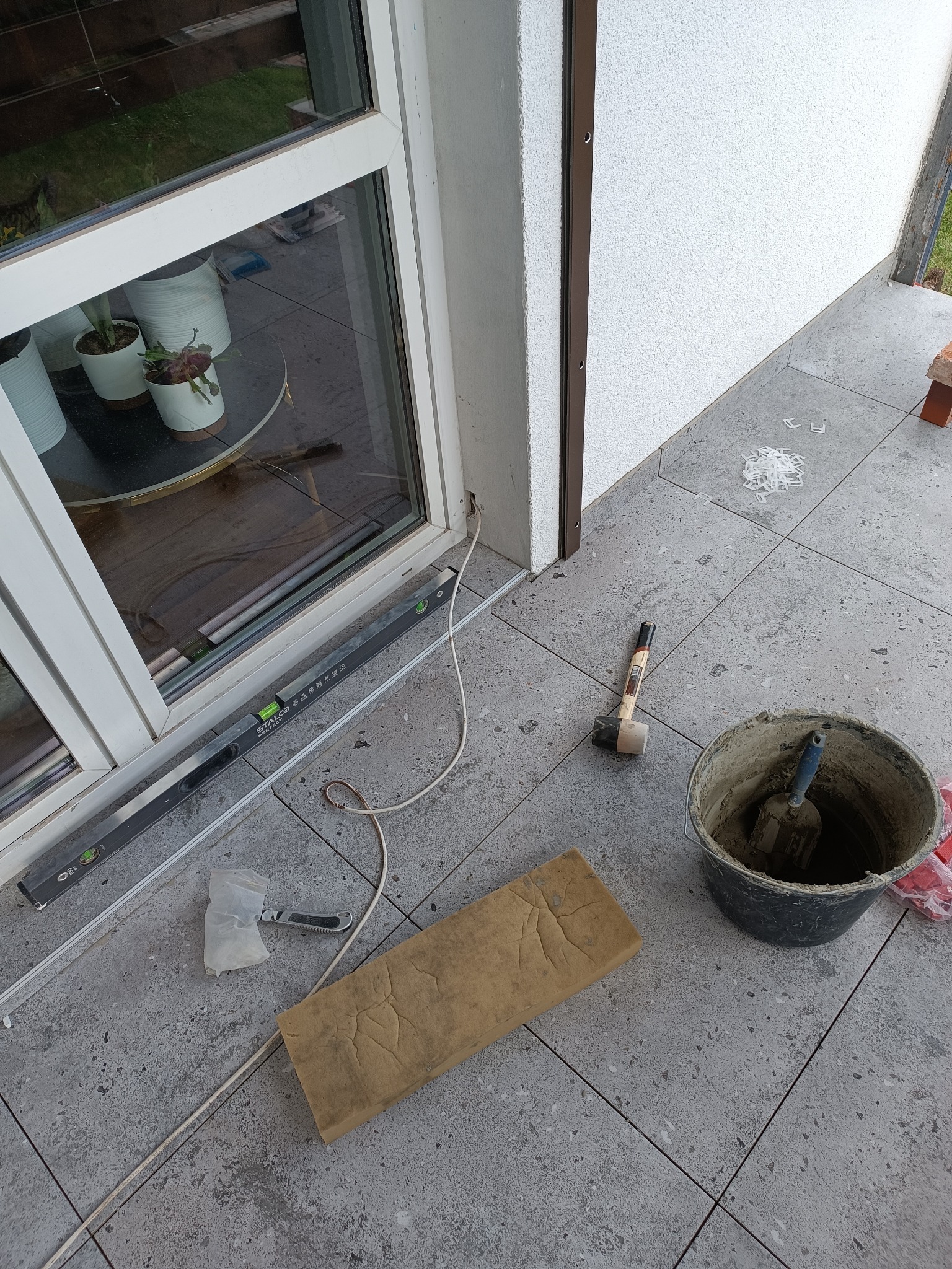 Szare płytki podłogowe na tarasie w trakcie układania, widoczne narzędzia: poziomica, gumowy młotek, wiadro z zaprawą, szpachelka i krzyżyki dystansowe.