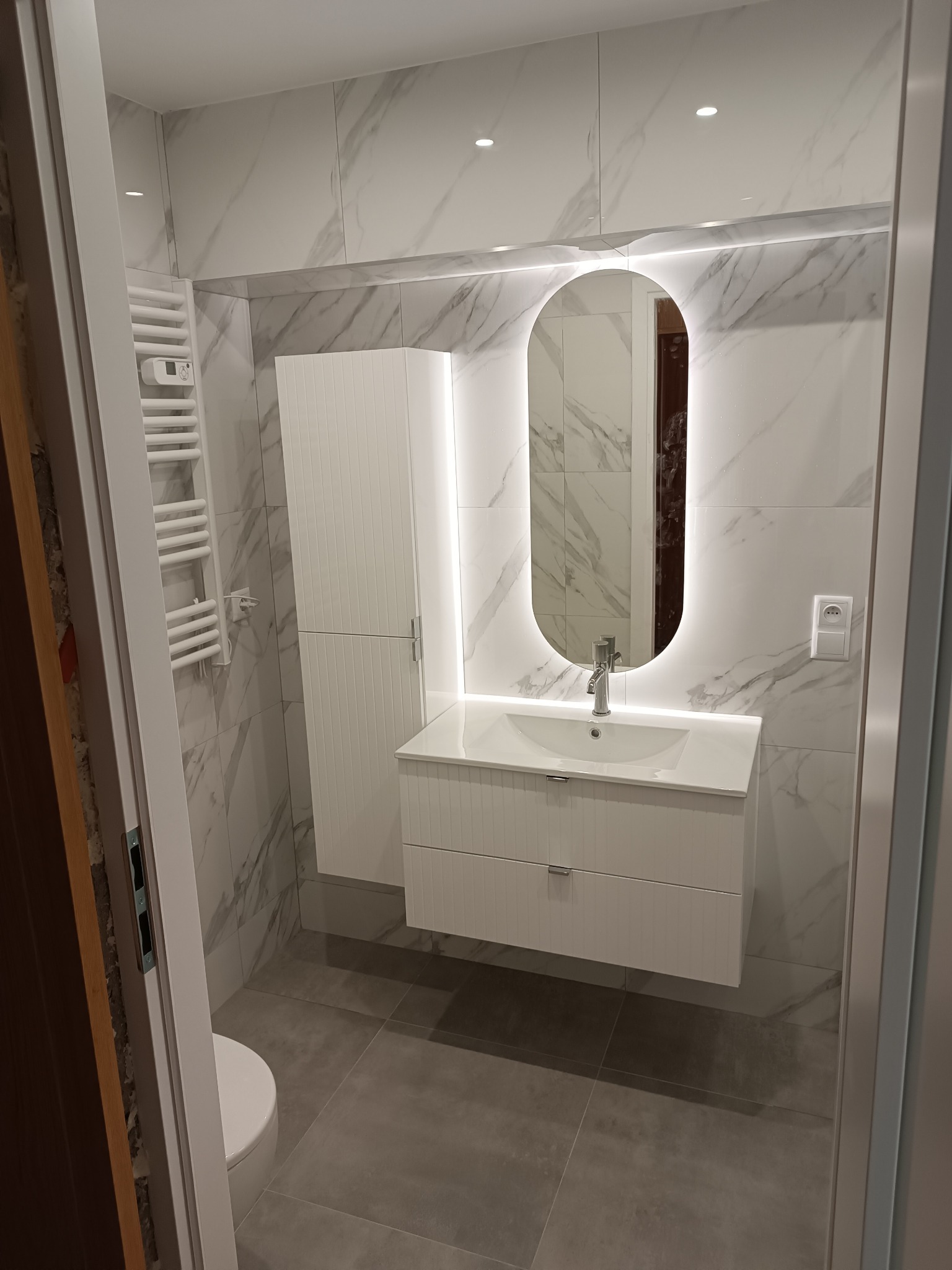 Nowoczesna łazienka z marmurowymi płytkami, podświetlanym lustrem i białymi meblami. Widoczny grzejnik drabinkowy i szara podłoga. Ujęcie z perspektywy wejścia.
