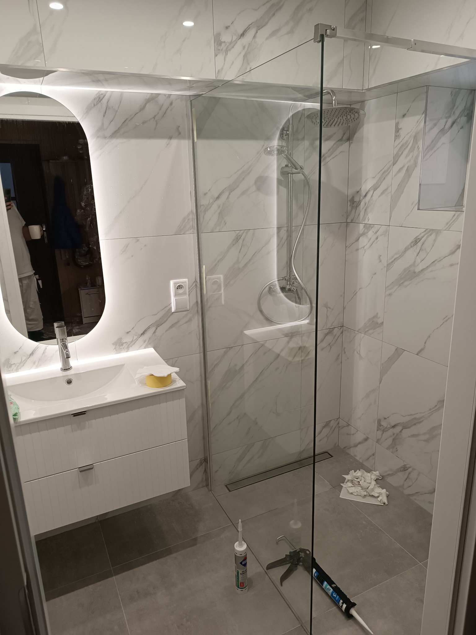 Nowoczesna łazienka z kabiną prysznicową typu walk-in, umywalką z szafką i lustrem z podświetleniem LED. Płytki imitujące marmur na ścianach i szara podłoga.