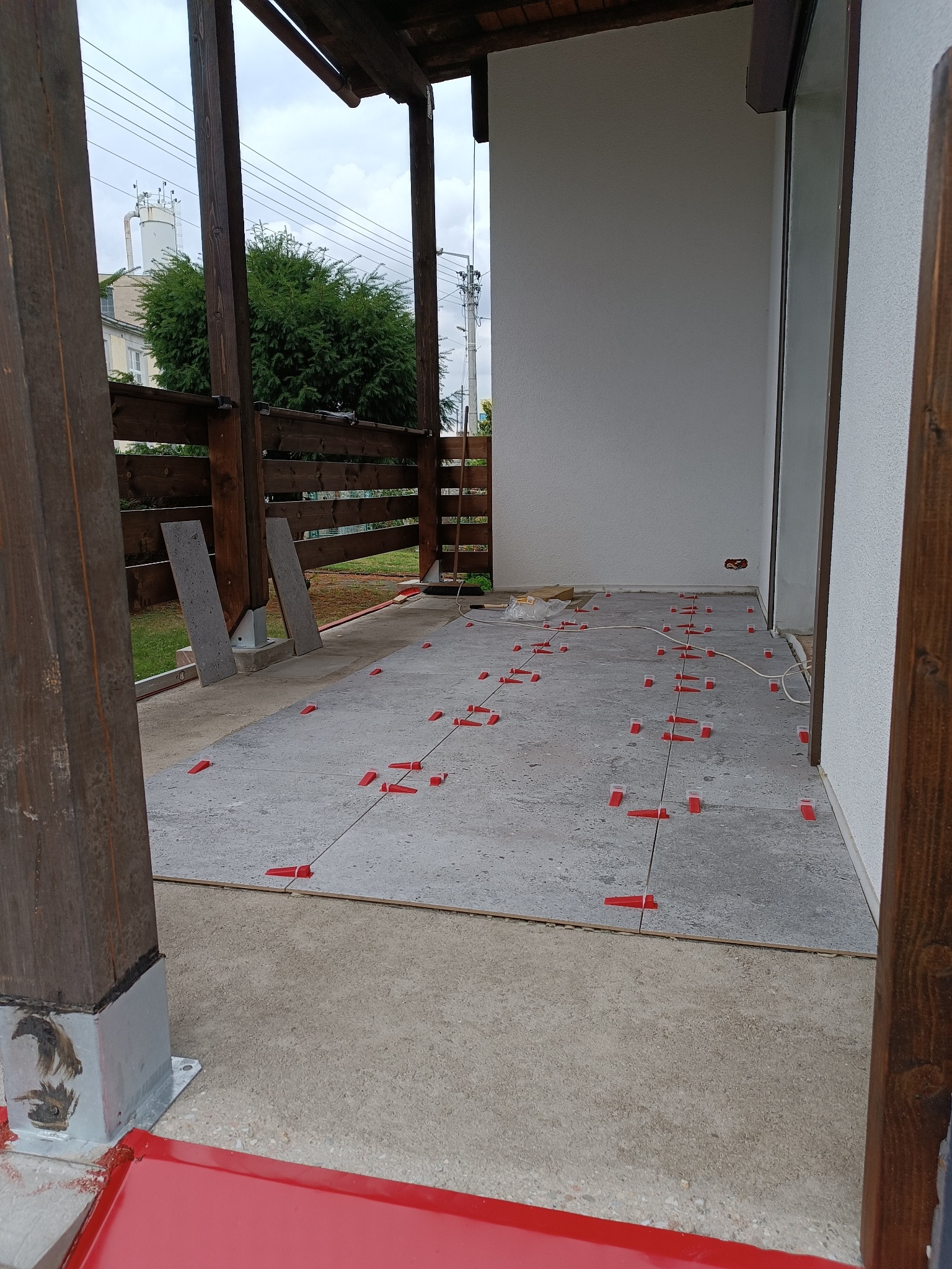 Układanie szarych płytek na tarasie z użyciem czerwonych klinów poziomujących. Widoczne drewniane elementy konstrukcji zadaszenia i fragment budynku w tle.