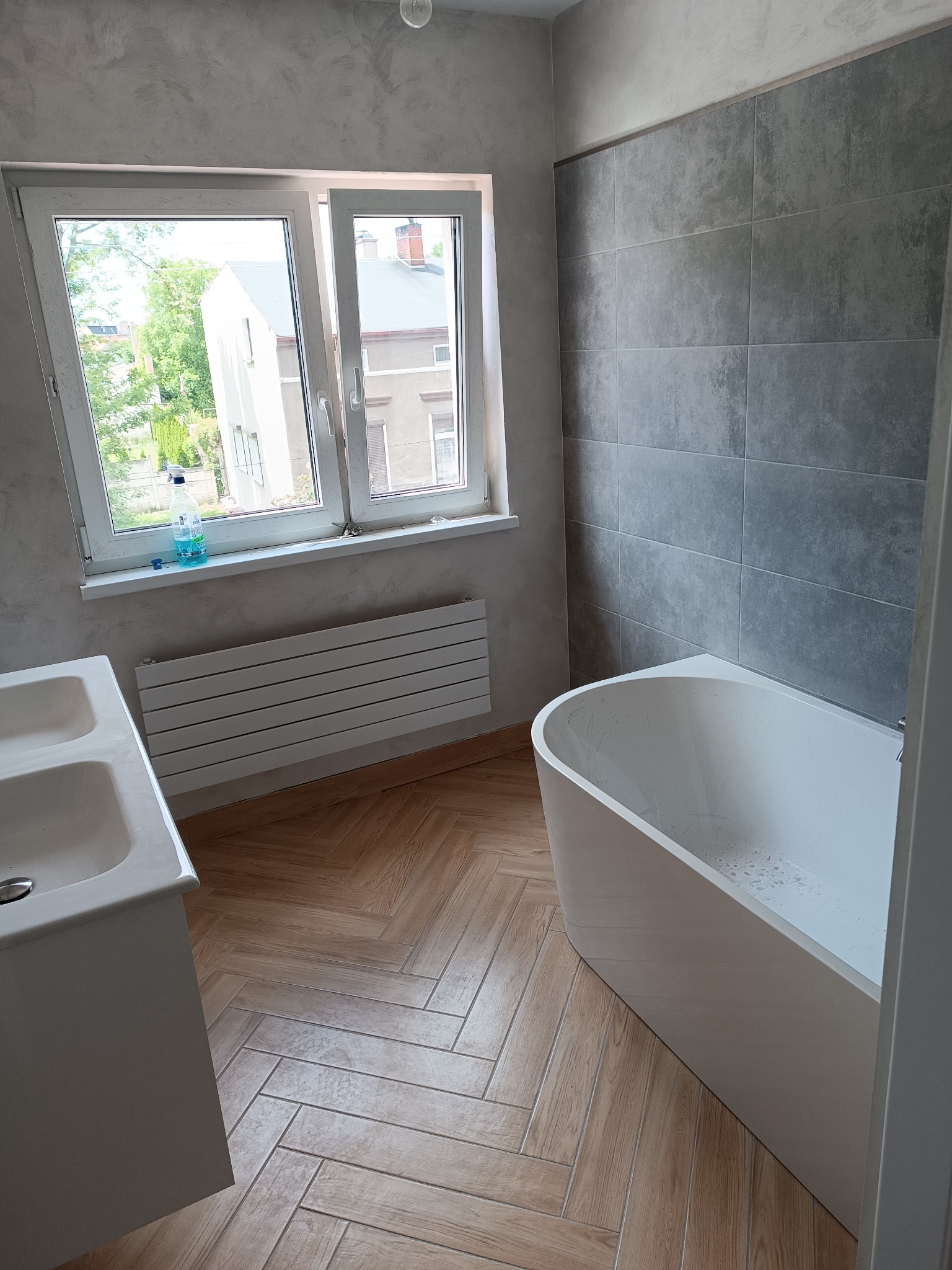 Nowoczesna łazienka po remoncie: szare płytki, drewnopodobna podłoga w jodełkę, biała wanna i umywalka. Widok na okno z zielenią na zewnątrz.