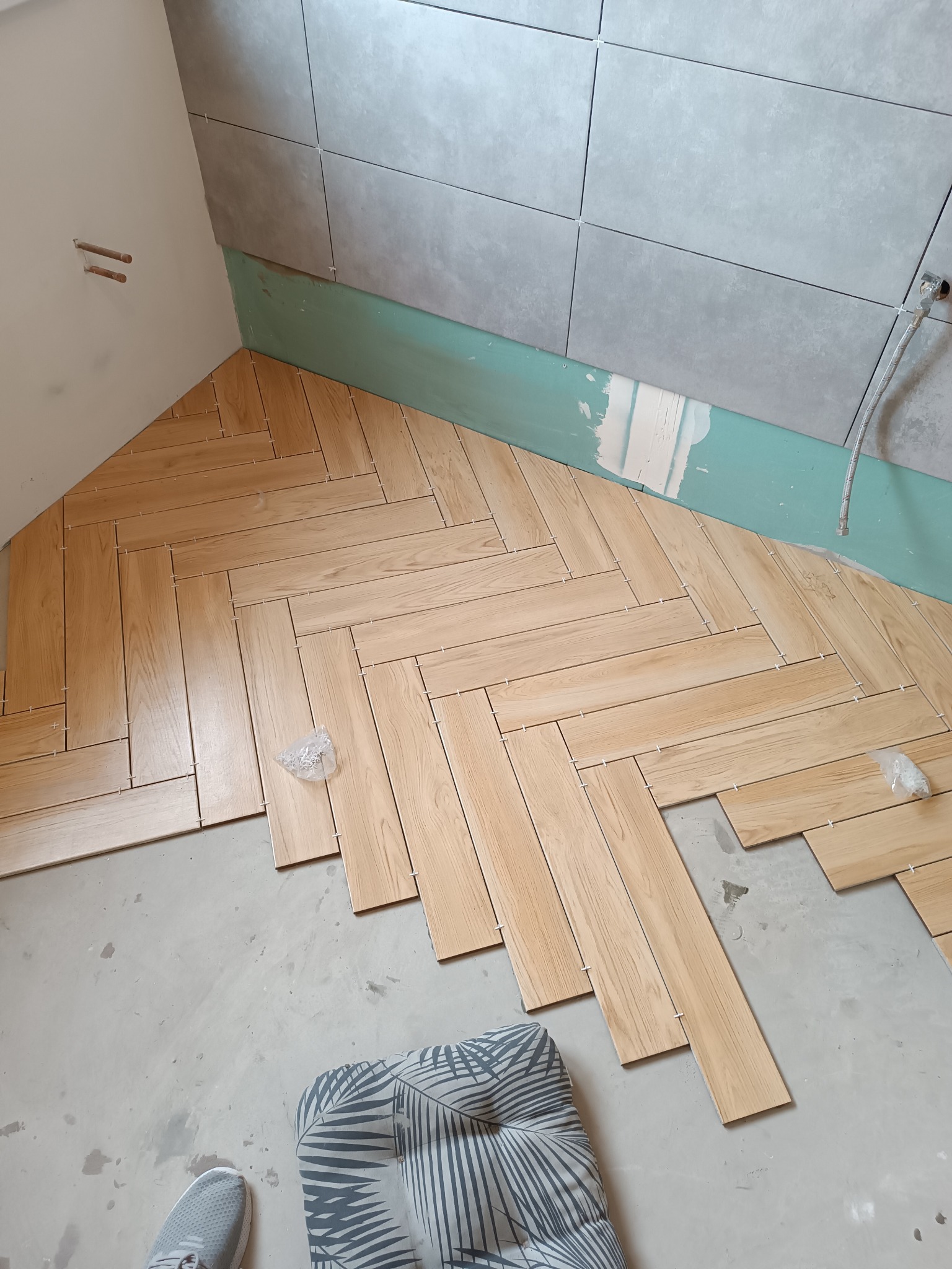 Układanie drewnopodobnych płytek w jodełkę na podłodze w łazience. Widoczne płytki na ścianie i akcesoria glazurnicze. Częściowo ukończony etap prac.