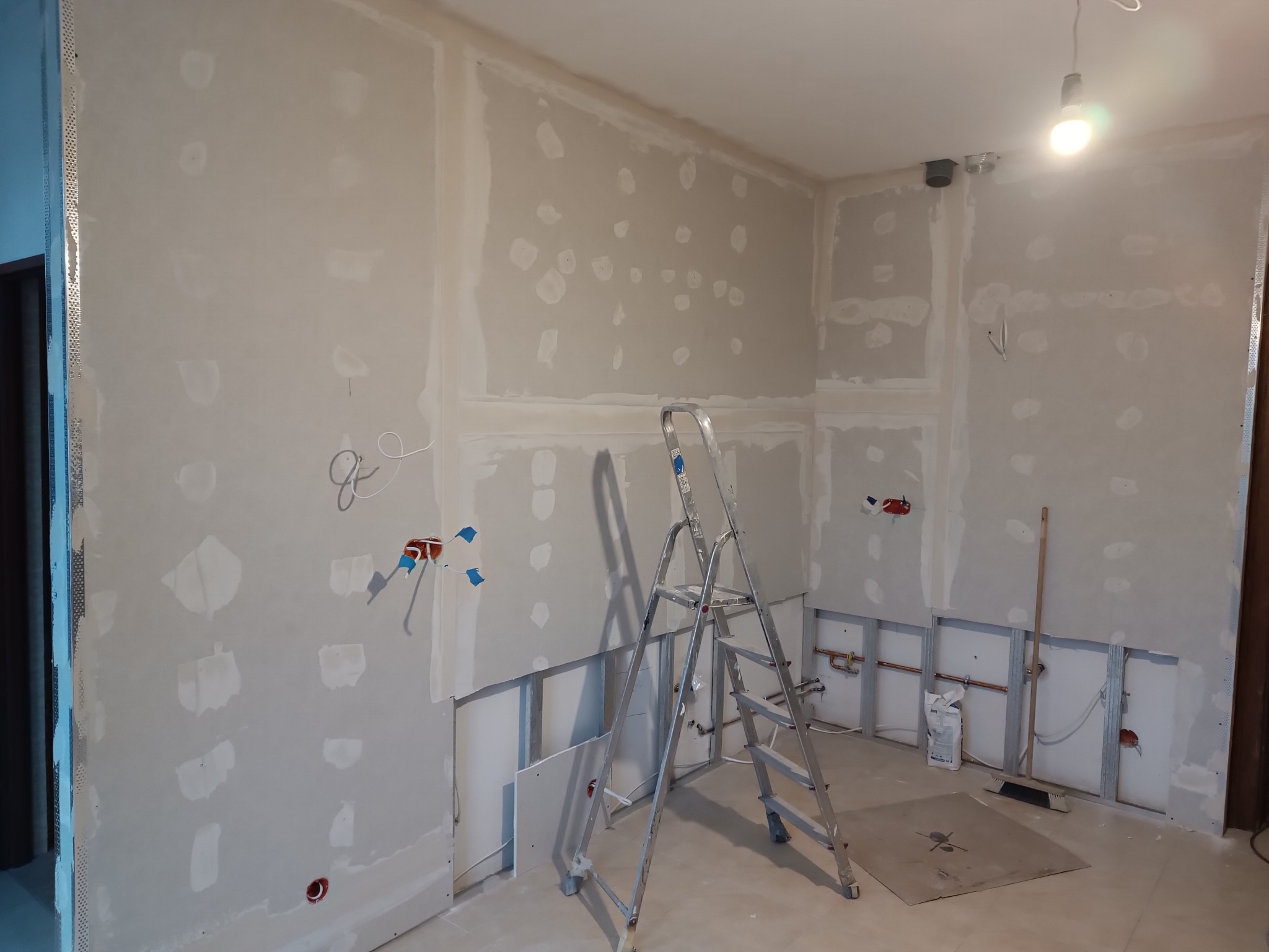 Wnętrze w trakcie prac wykończeniowych: ściany z płyt gipsowo-kartonowych, widoczne instalacje, drabina. Oświetlenie sufitowe. Etap szpachlowania i przygotowania do malowania.