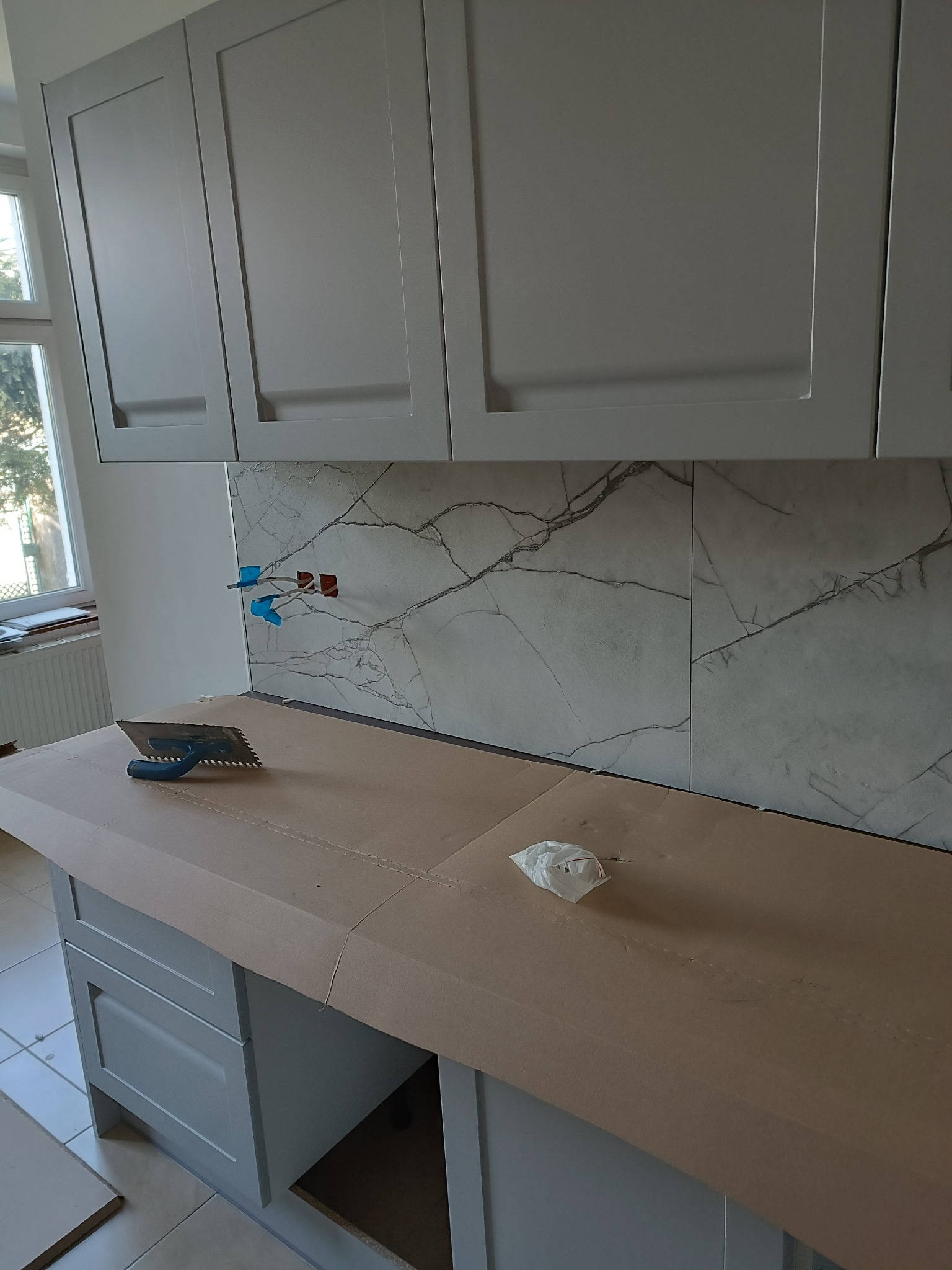 Montaż płytek imitujących marmur na ścianie nad blatem kuchennym. Widoczna szpachelka i zabezpieczony blat. Szare szafki kuchenne w tle.