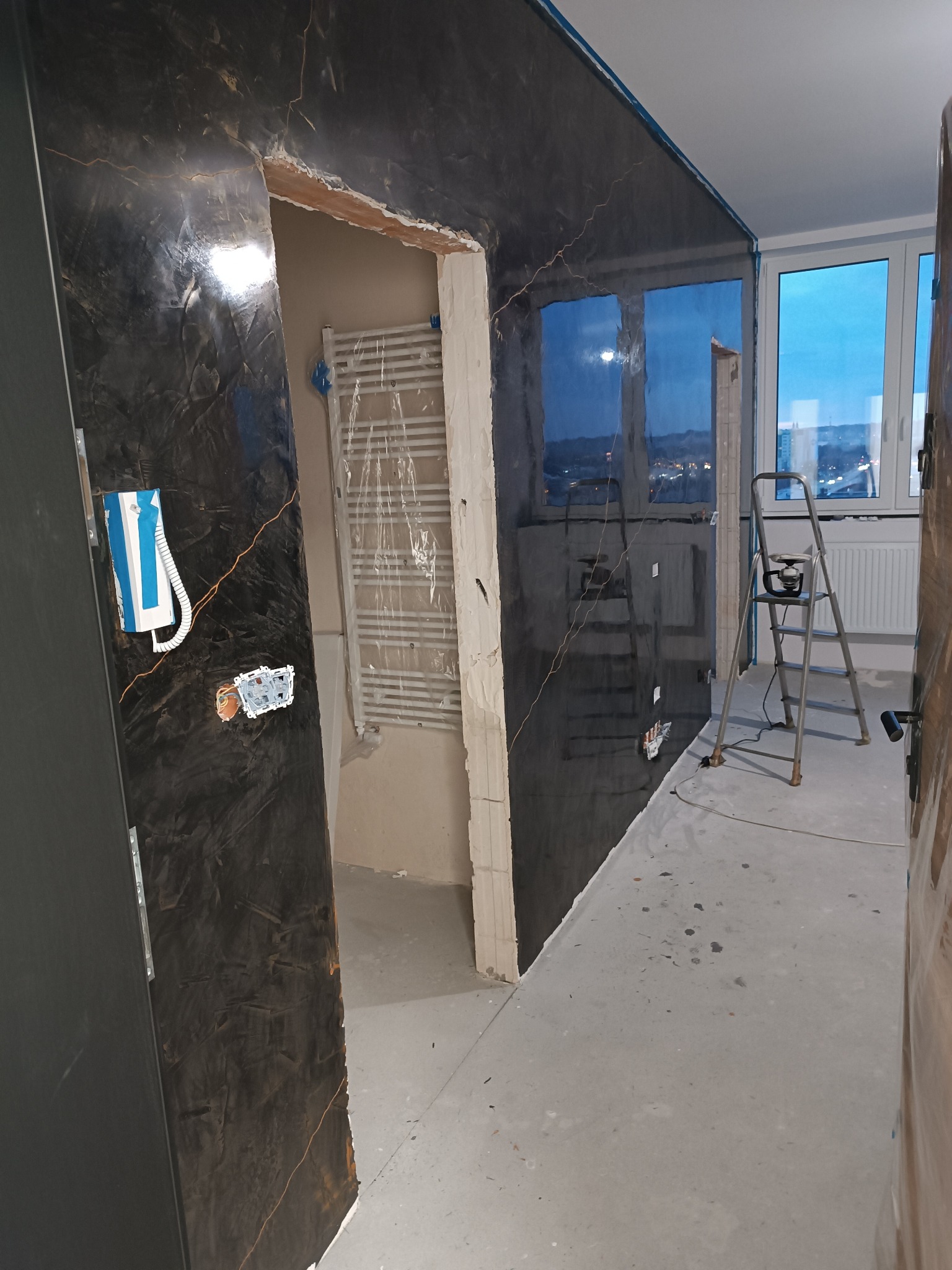 Ciemna ściana z efektem marmuru w trakcie wykańczania, widoczny grzejnik i drabina, okno z widokiem na miasto w tle. Fragment instalacji elektrycznej na ścianie.