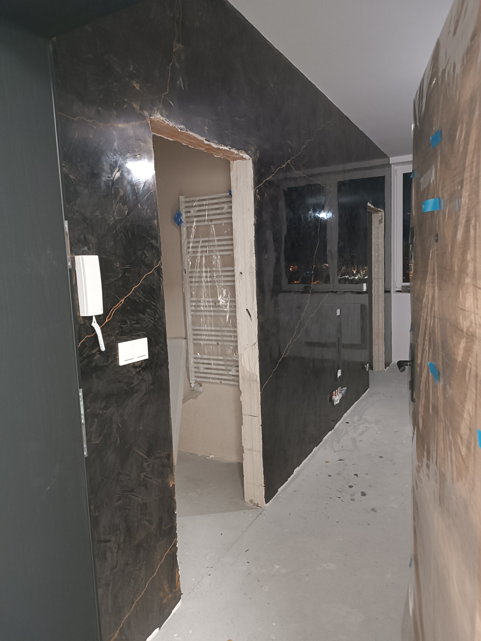 Wykończenie ścian w korytarzu z efektem marmuru, widoczny grzejnik i fragment okna. Ściany w ciemnych odcieniach z jasnymi żyłkami, surowy stan wykończenia.