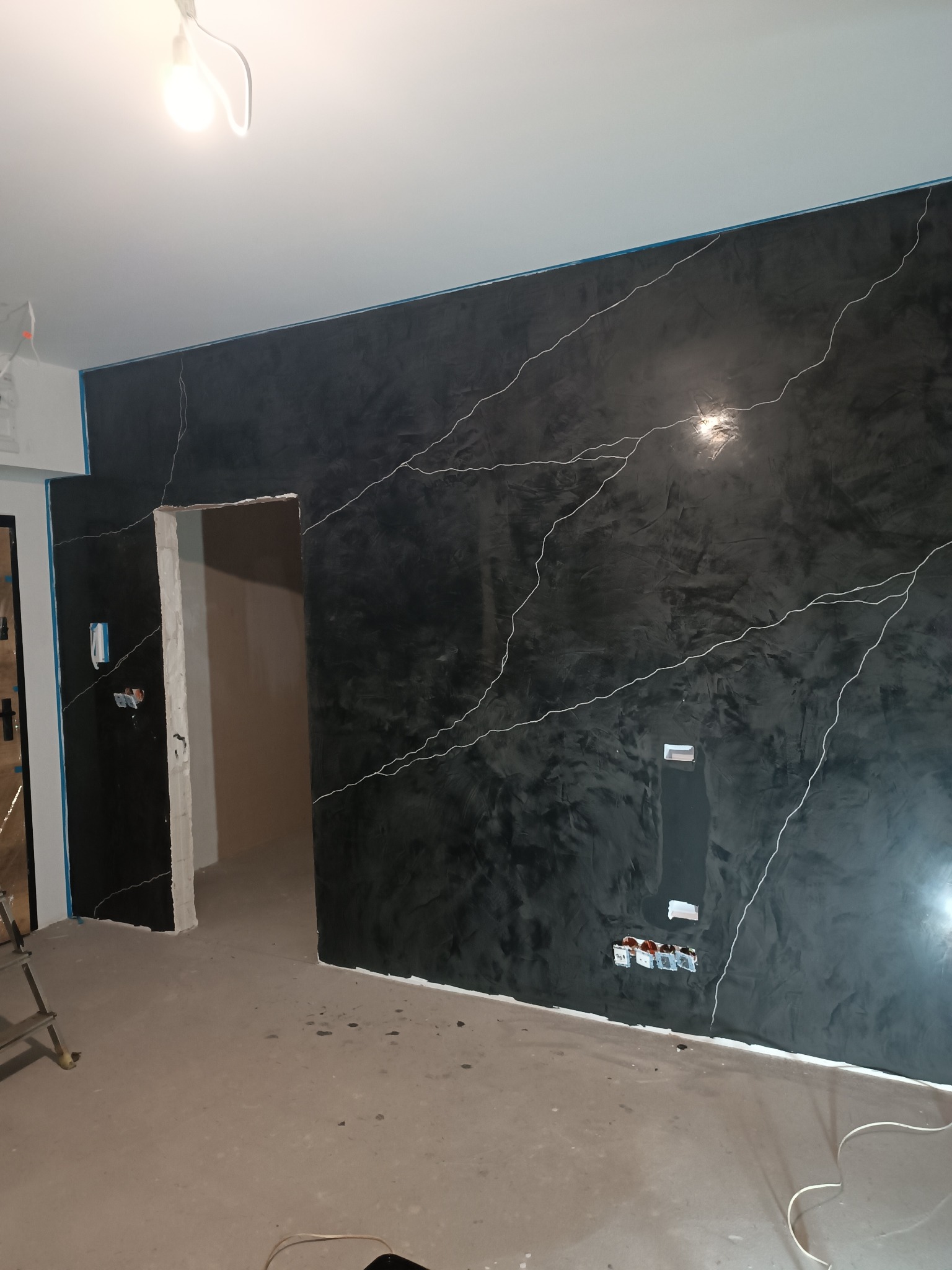 Ściana w pokoju po malowaniu dekoracyjnym w odcieniach czerni z białymi żyłkami, widoczne puszki elektryczne i fragment drabiny.