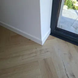 Jasne panele podłogowe ułożone przy białej ścianie z listwą przypodłogową i czarną ramą okienną. Widok na zieleń za oknem.