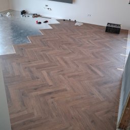 Wall-print - Częściowo ułożona podłoga z paneli w jodełkę, widoczne narzędzia i podkład. Szara ściana z wnęką w tle. Etap prac wykończeniowych w nowym wnętrzu.