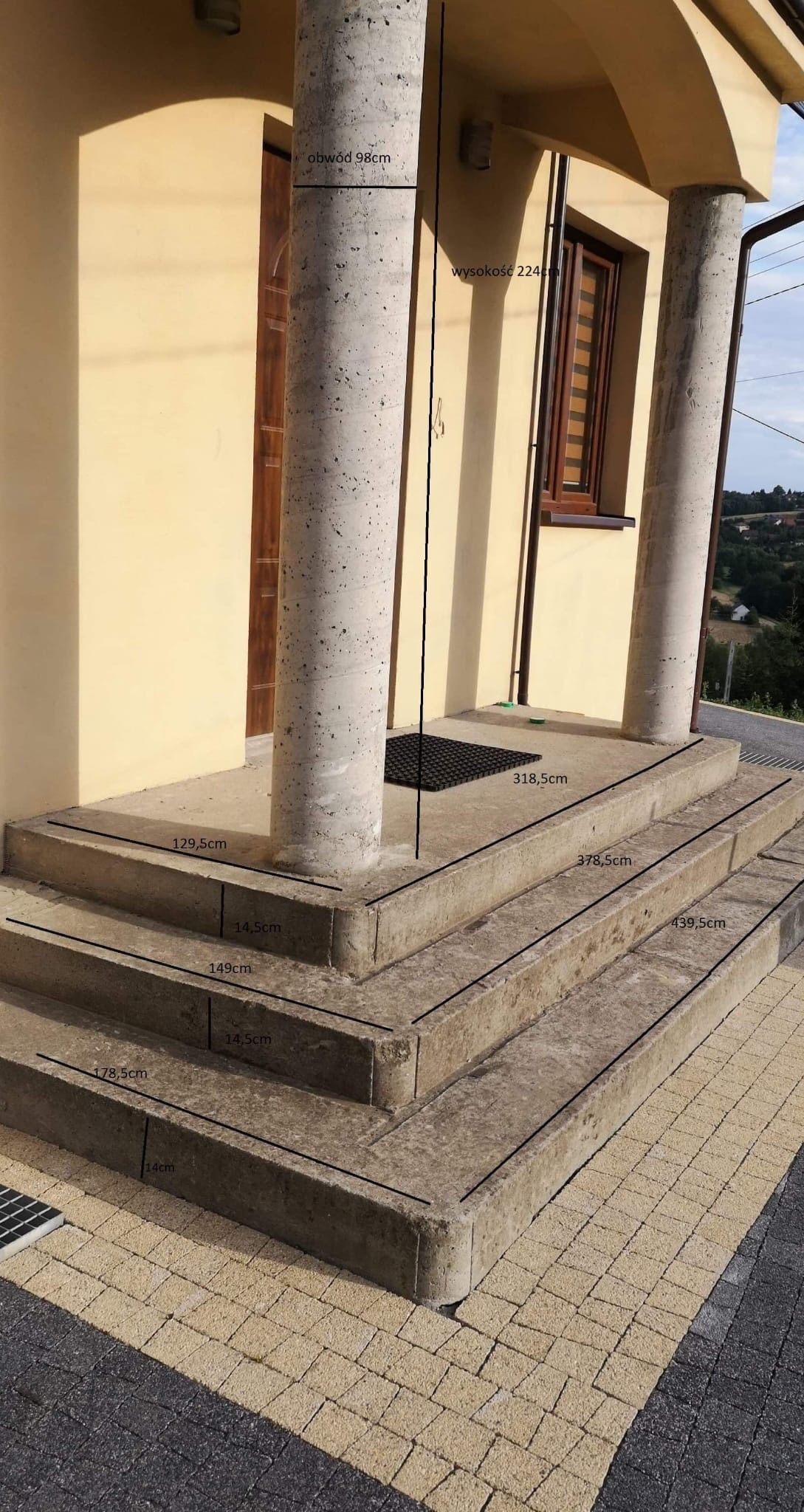 Wejście do budynku z trzema betonowymi schodami, dwoma kolumnami z szarego betonu, brązowymi drzwiami i oknem, z naniesionymi liniami z wymiarami.