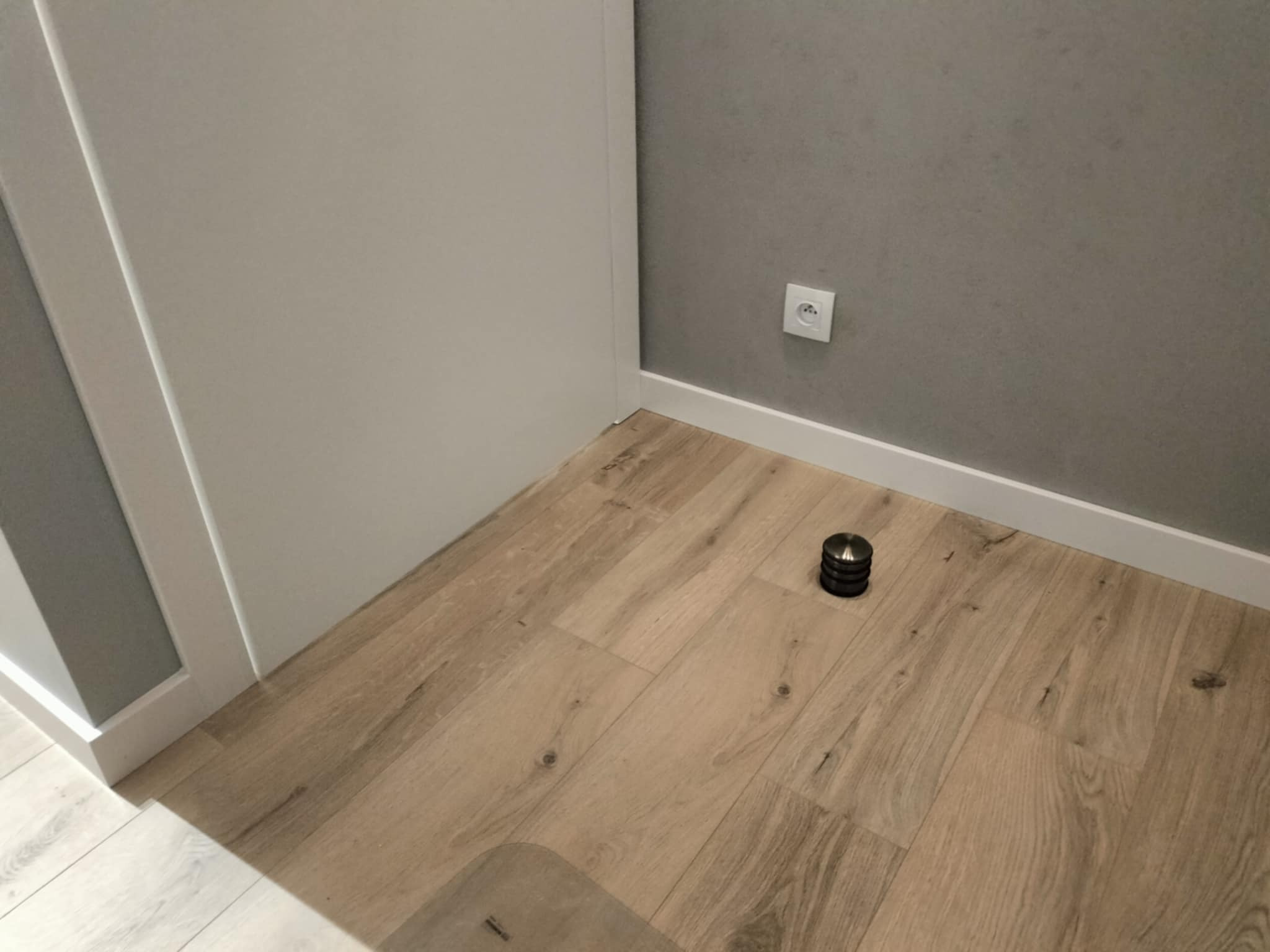Ułożone panele podłogowe imitujące drewno, widoczna listwa przypodłogowa, szara ściana z gniazdkiem elektrycznym oraz ogranicznik do drzwi w kolorze czarnym.