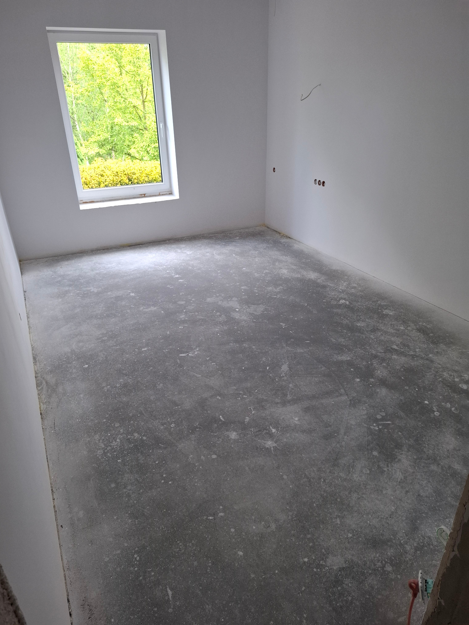 Przygotowane podłoże w pustym, jasnym pokoju z oknem, gotowe do położenia paneli. Wyrównana wylewka, widoczne ślady szlifowania. Gniazdka elektryczne na ścianie.