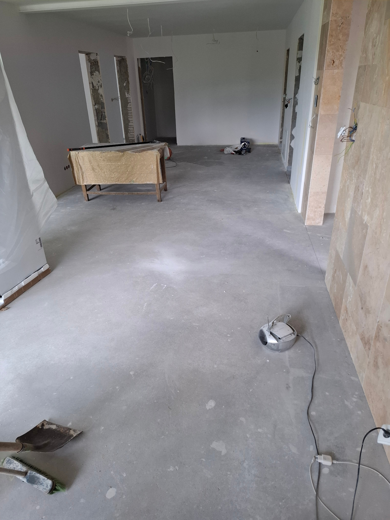 Wnętrze w trakcie remontu: surowy beton na podłodze, częściowo wykończone ściany, narzędzia budowlane. Widoczne przygotowania do dalszych prac wykończeniowych.