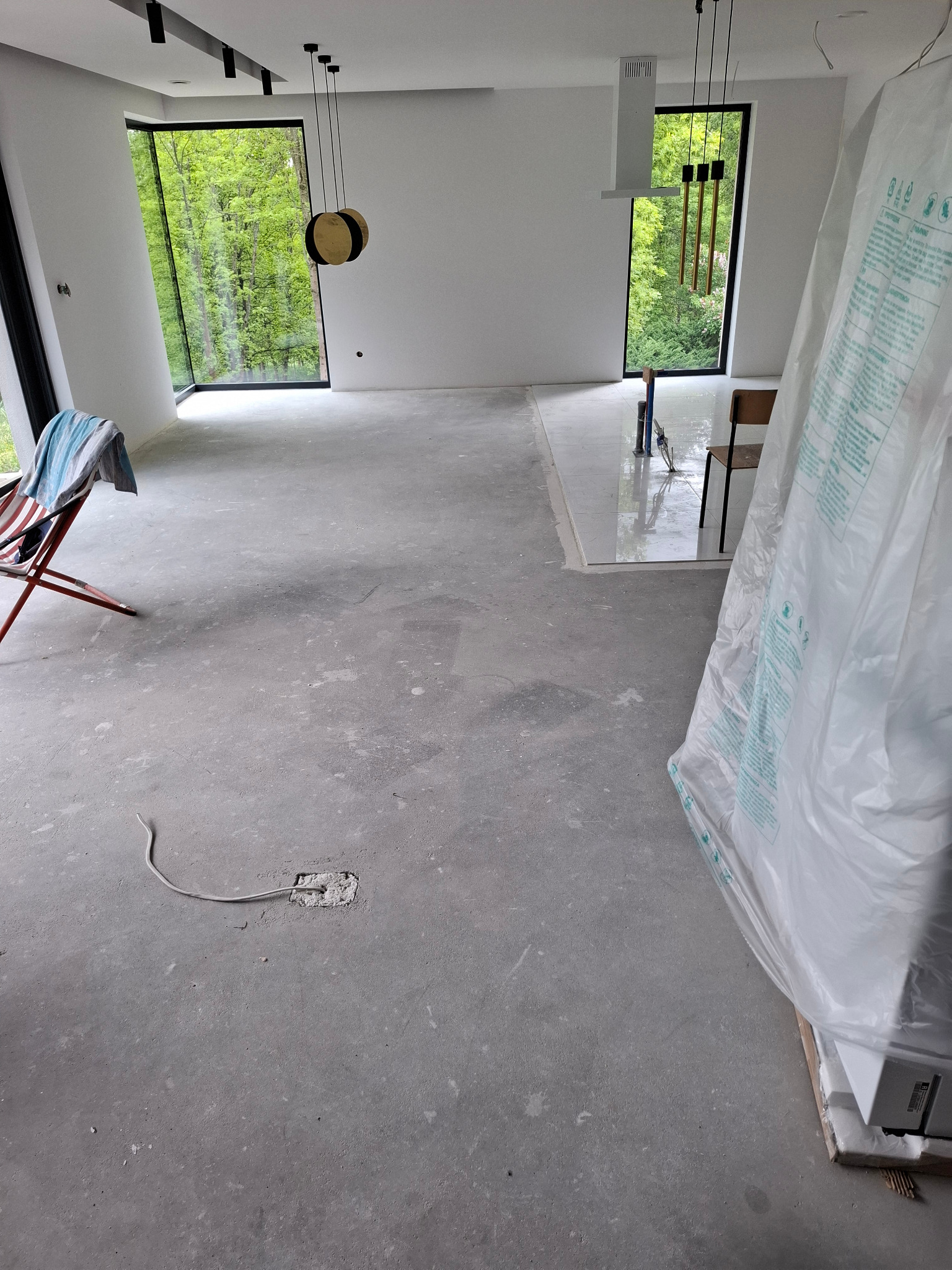 Wnętrze w trakcie remontu: szare podłoże, widoczne okna z zielonym tłem, elementy instalacji i sprzęt zabezpieczony folią. Prace wykończeniowe w nowoczesnym stylu.