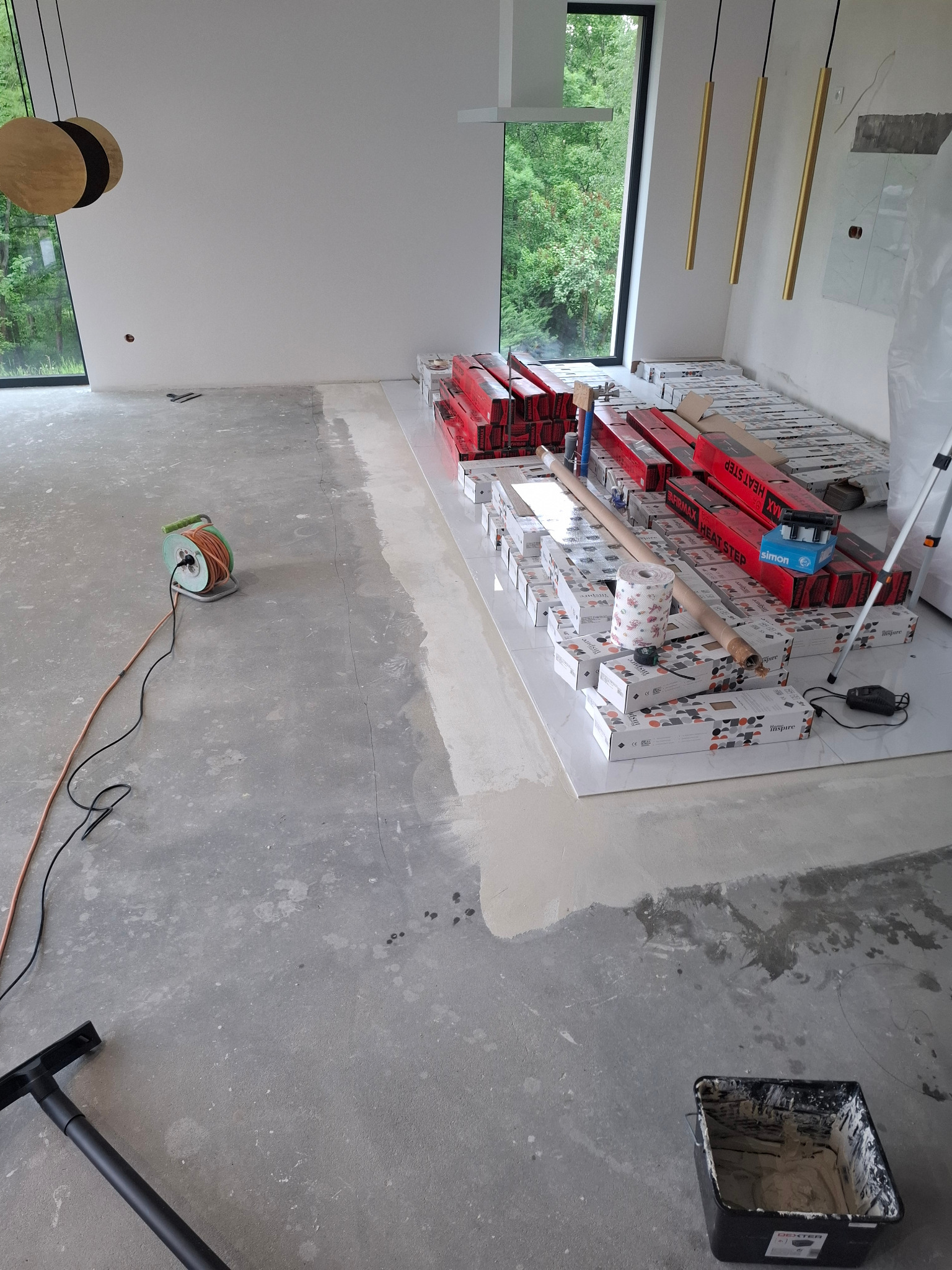 Przygotowanie do położenia podłogi: wyrównana wylewka, panele i akcesoria ułożone na białych płytkach. Widok na okno z zielenią w tle.