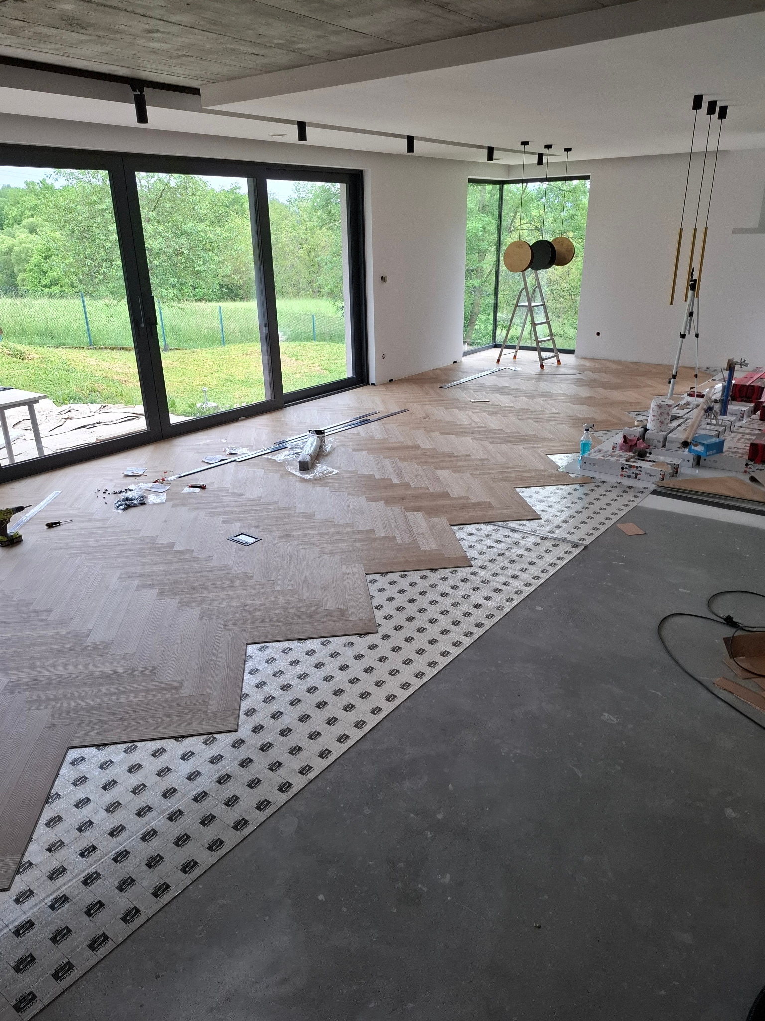 Układanie parkietu w jodełkę francuską, widoczne narzędzia i podkład. Duże okna z widokiem na zieleń, surowy betonowy sufit.