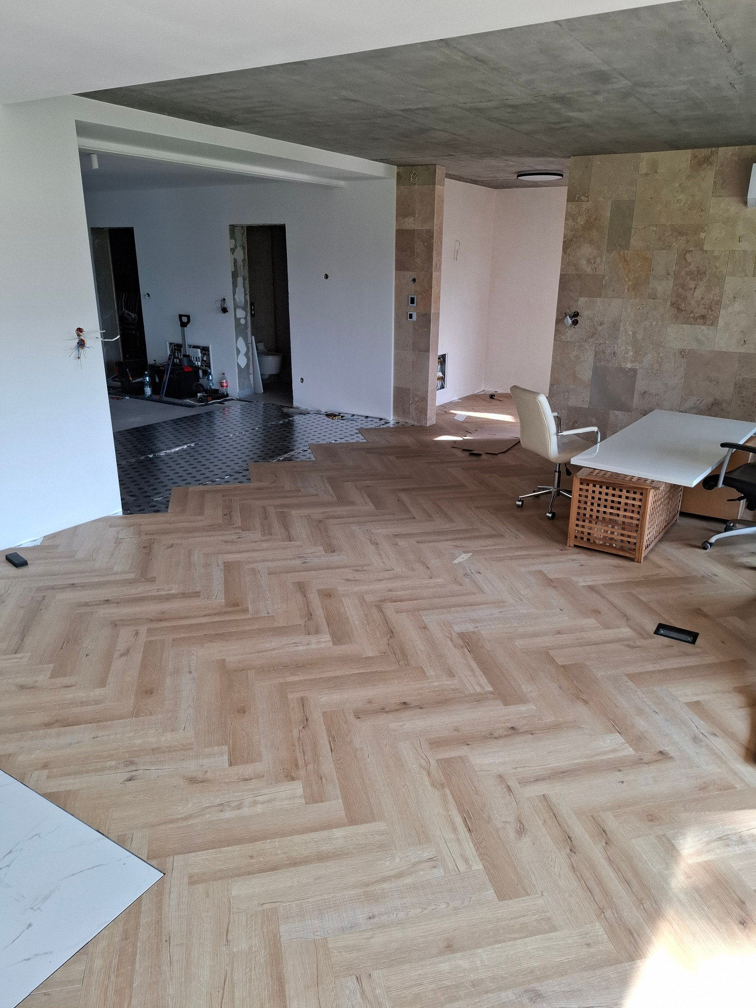 Układanie jasnych paneli podłogowych w jodełkę w przestronnym wnętrzu. Częściowo widoczna podkładka izolacyjna i płytki. W tle biuro z kamienną ścianą.