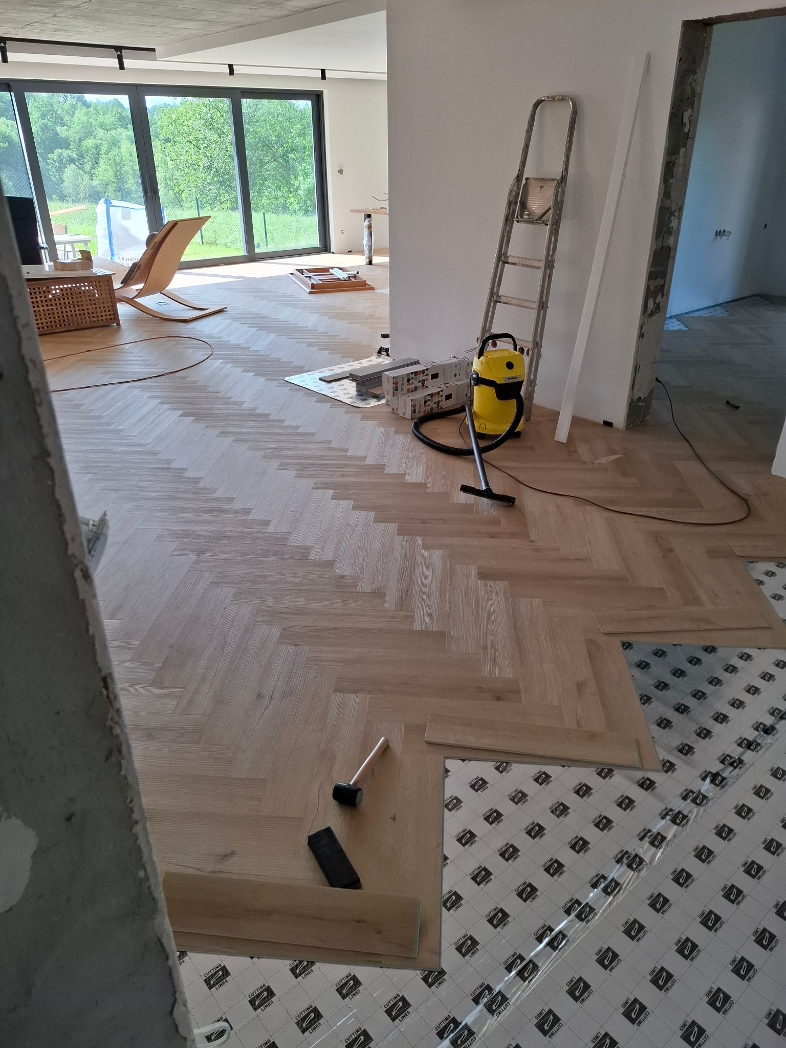 Układanie parkietu w jodełkę, widoczne narzędzia i odkurzacz. Część podłogi zabezpieczona folią. Duże okno z widokiem na zieleń w tle.