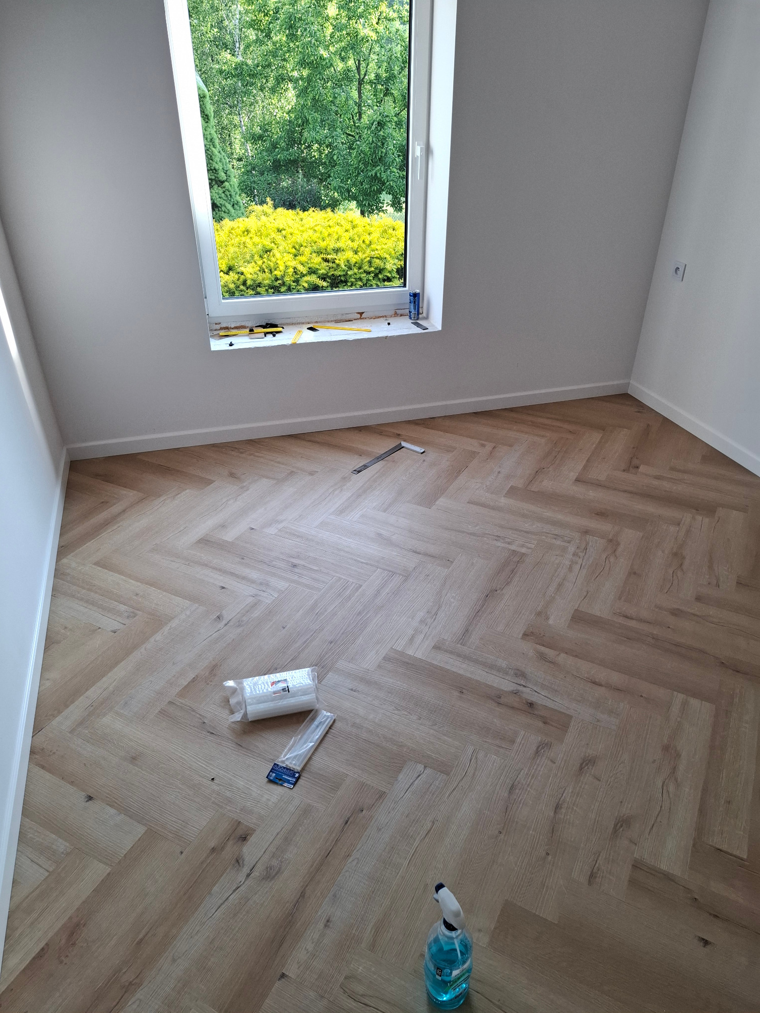 Jasny parkiet ułożony w jodełkę w pokoju z białymi ścianami i oknem z widokiem na zieleń. Na podłodze narzędzia i akcesoria do montażu podłogi.