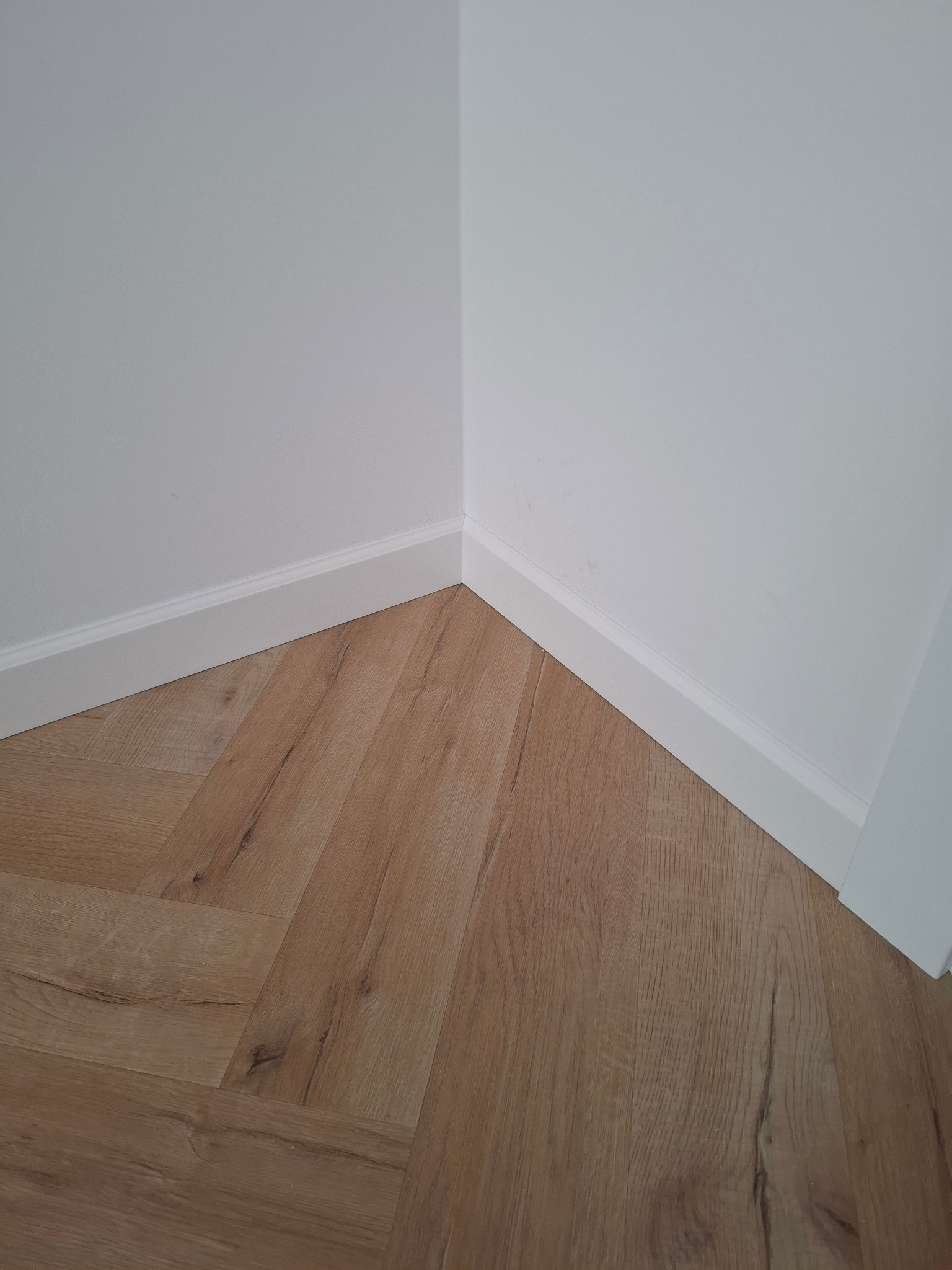 Jasne panele ułożone w jodełkę francuską przy białej ścianie z listwami przypodłogowymi. Widok na róg pomieszczenia z bliska.