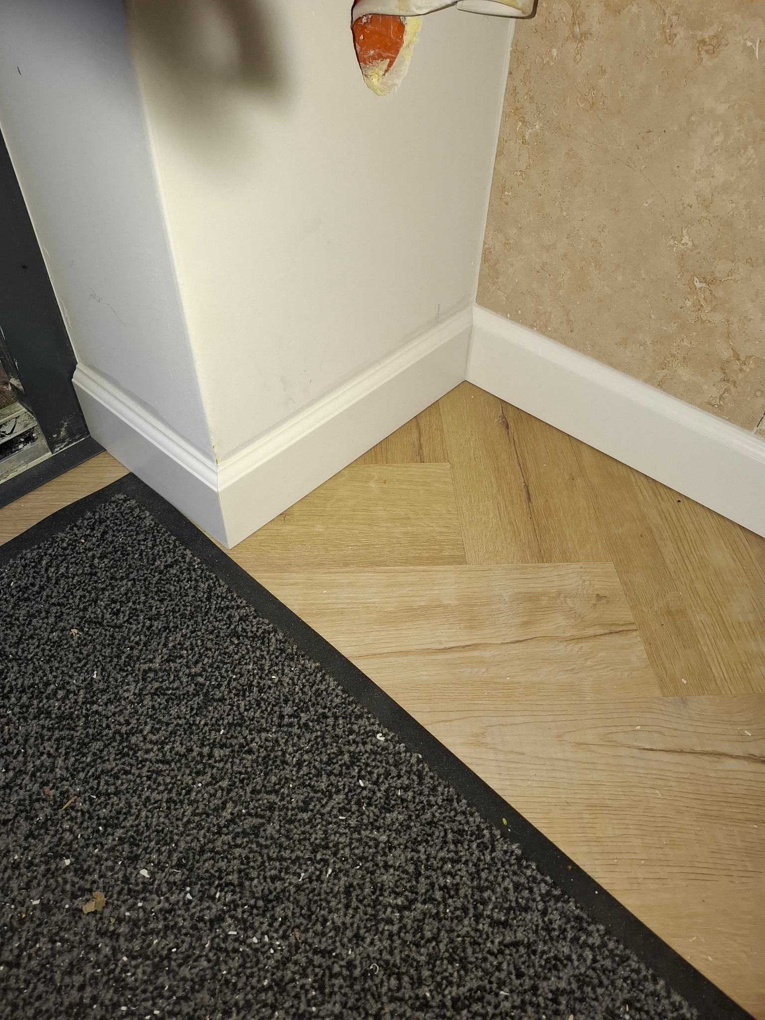 Fragment wykończonego wnętrza: panele podłogowe ułożone w jodełkę, białe listwy przypodłogowe, fragment ściany z beżową okładziną i szara wycieraczka.