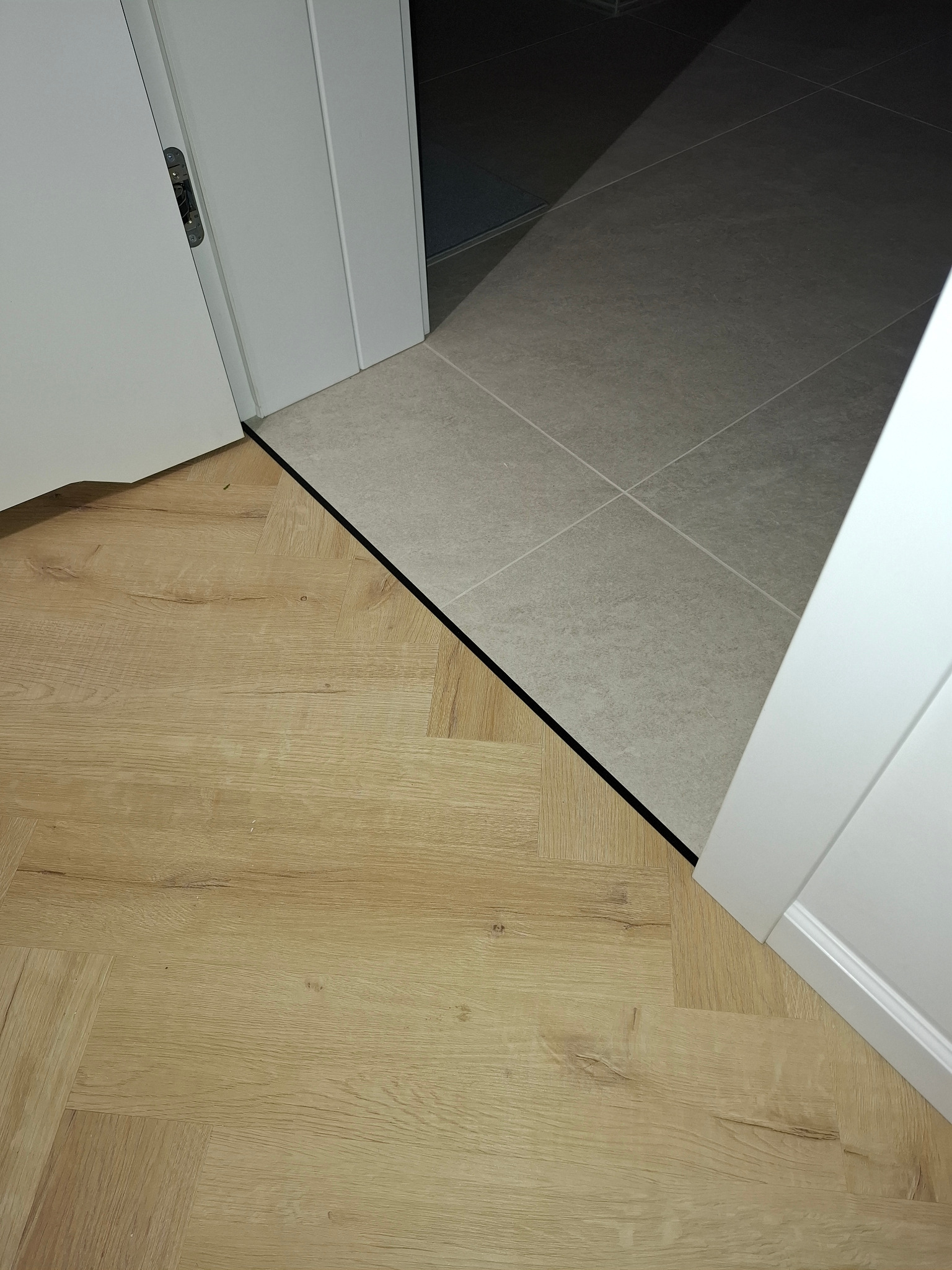 Połączenie parkietu w jodełkę i szarych płytek w progu drzwi. Widoczne białe ościeżnice i czarny pasek dylatacyjny. Kontrast materiałów i kolorów.