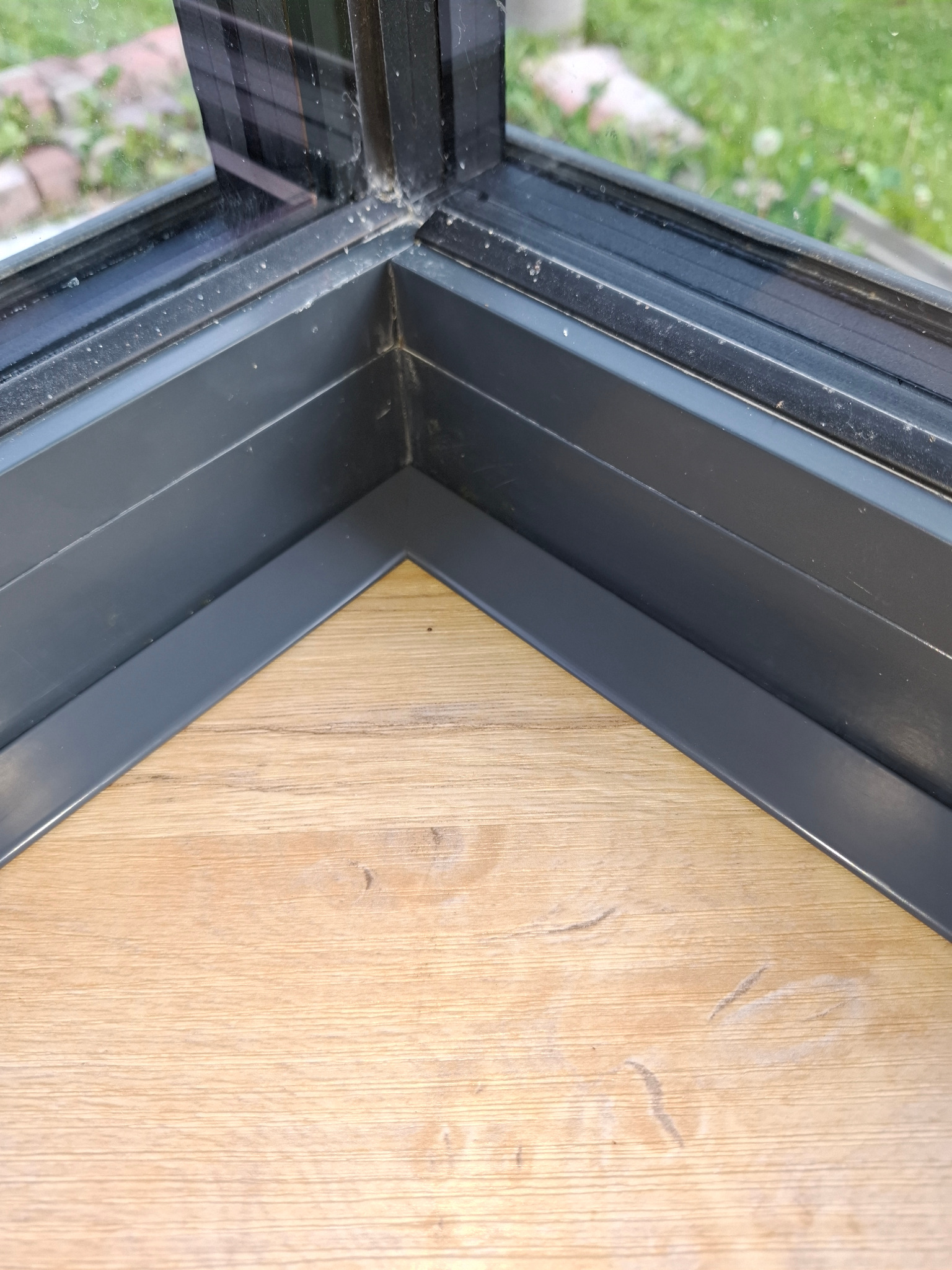 Detal ukończonego rogu podłogi z paneli drewnopodobnych, przylegającej do ramy okiennej z widokiem na zieleń za oknem. Precyzyjne wykończenie.