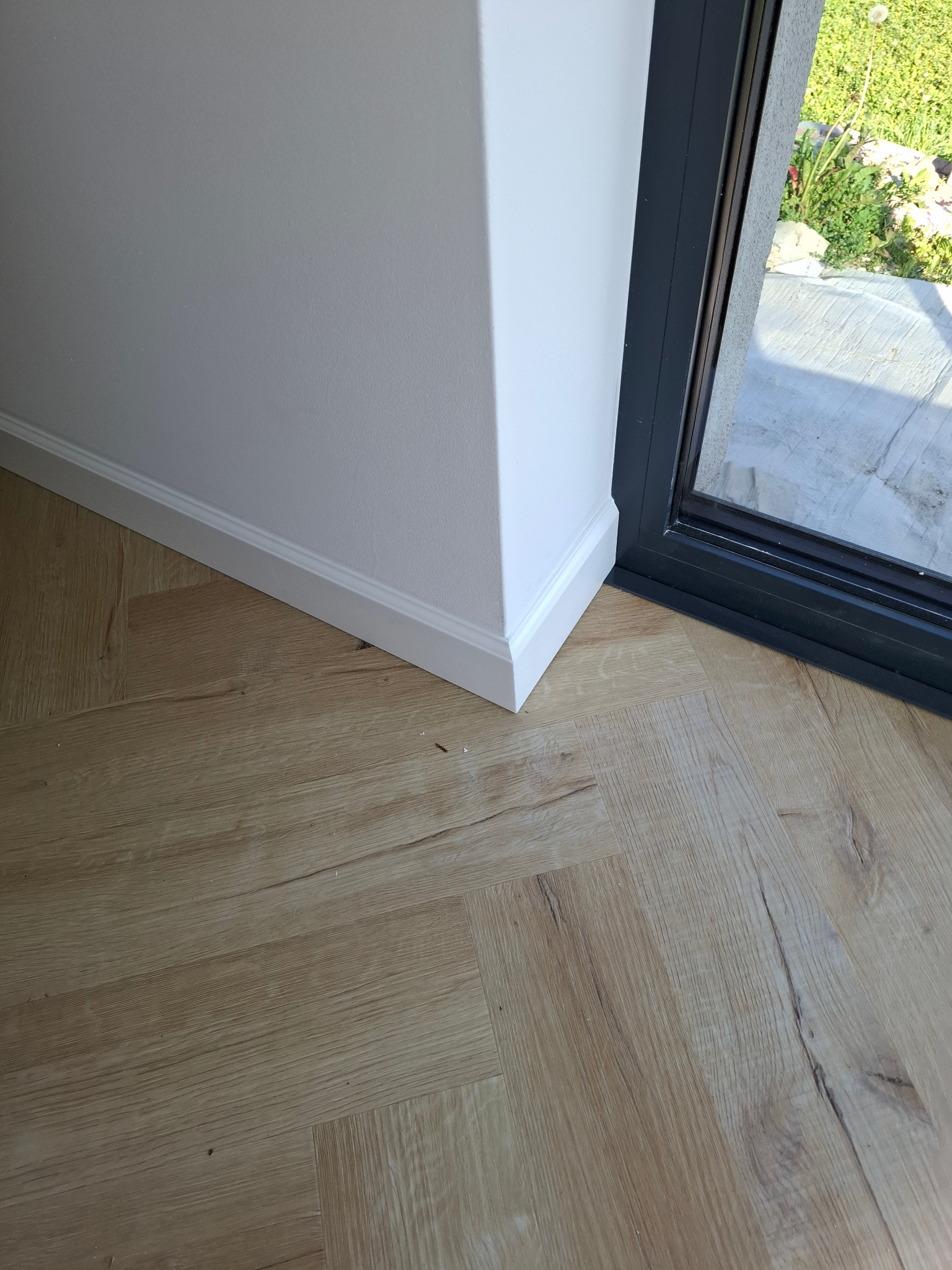 Jasne panele podłogowe ułożone przy białej ścianie z listwą przypodłogową i czarną ramą okienną. Widok na zieleń za oknem.