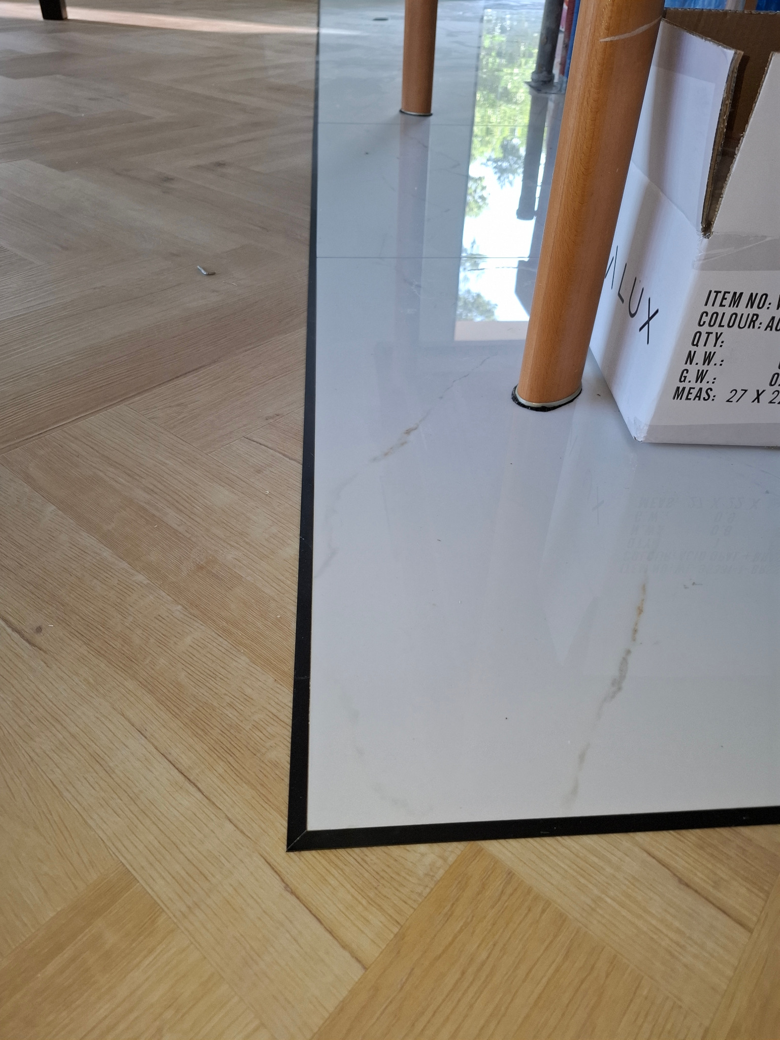 Połączenie parkietu i białych płytek podłogowych z czarną listwą w Wieliczce. Widoczna kartonowa paczka i noga krzesła. Ujęcie z bliska, prezentujące detale wykończenia.