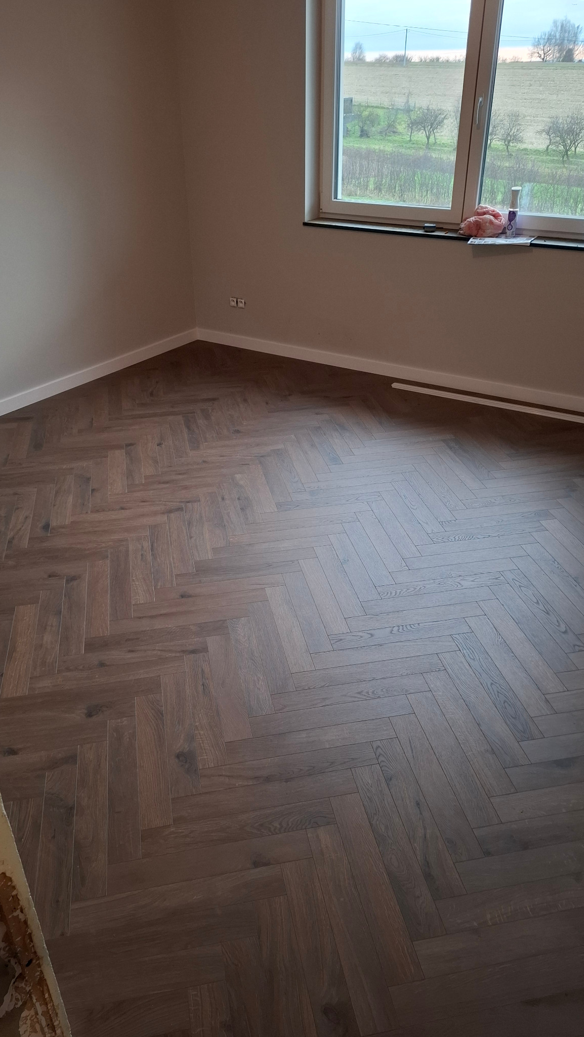 Ukończona podłoga z paneli ułożonych w jodełkę w jasnym pokoju z oknem na krajobraz. Widoczne listwy przypodłogowe i fragment niedokończonego wykończenia.