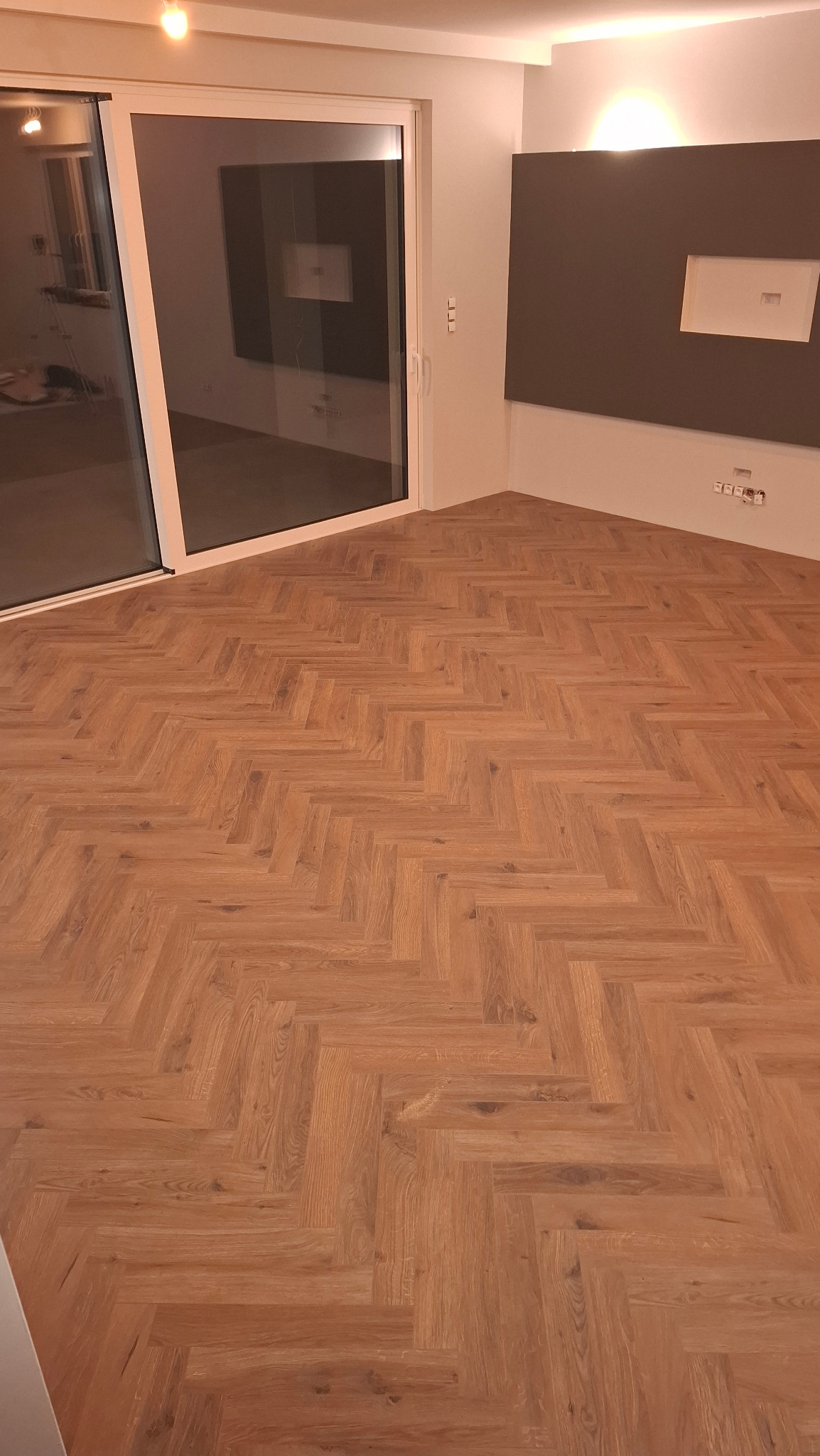 Jasny parkiet ułożony we wzór jodełki francuskiej w przestronnym pokoju z dużym oknem przesuwnym i szarą ścianą. Widoczne gniazdka elektryczne przy podłodze.