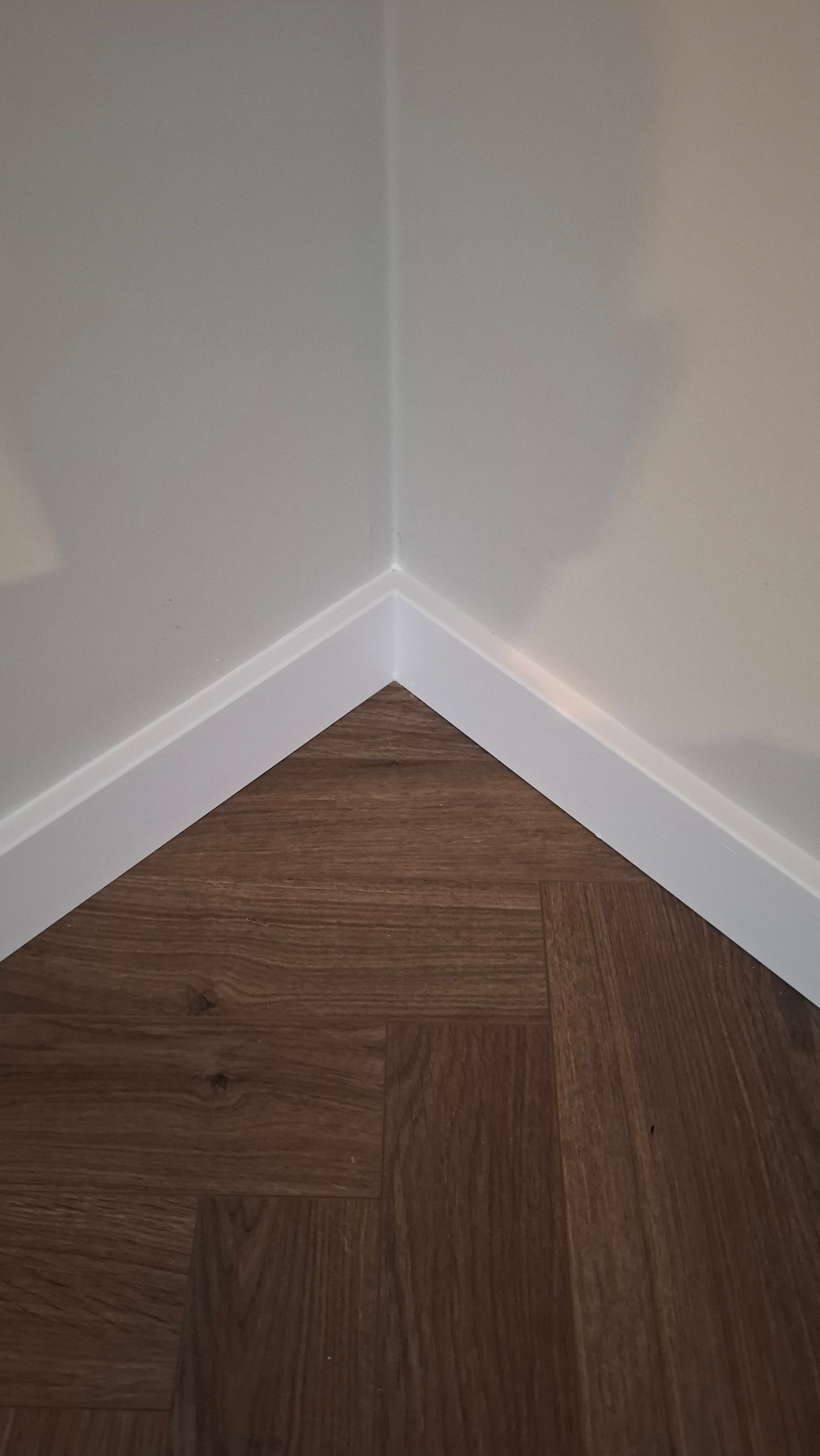 Ukończone połączenie podłogi z paneli w rogu pokoju, z białą listwą przypodłogową. Widoczny wzór paneli w jodełkę i gładka ściana.