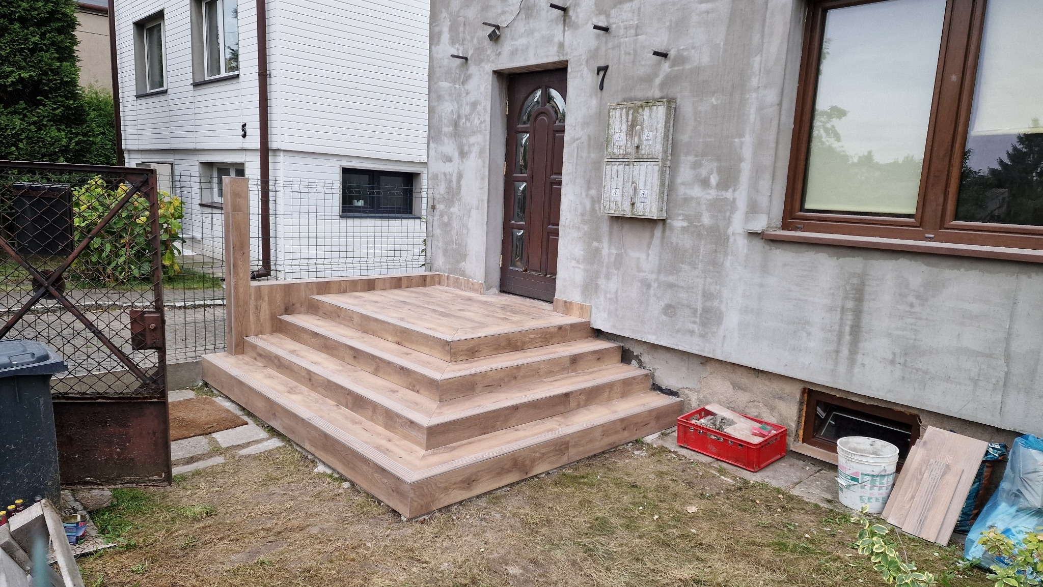Nowe schody wejściowe z drewnopodobnych płytek prowadzące do drzwi domu z numerem 7, widoczna skrzynka elektryczna, obok czerwony pojemnik z gruzem, na trawniku ślady budowy.