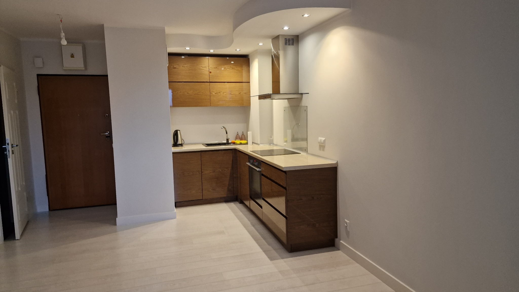 Nowoczesna kuchnia z drewnianymi szafkami i jasnym blatem, widok z perspektywy wejścia, z minimalistycznym okapem i oświetleniem punktowym.