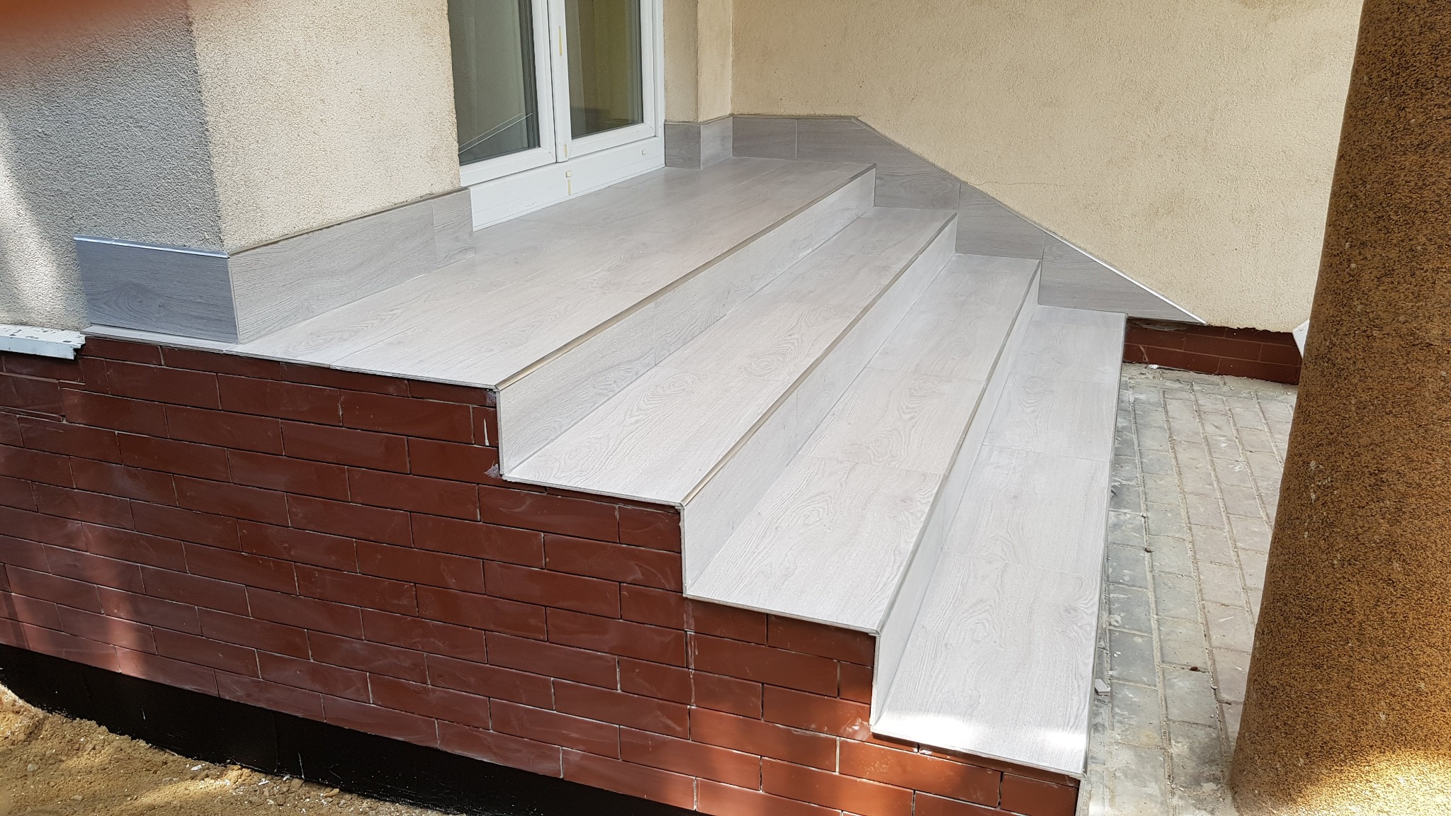 Wyłożone płytkami schody wejściowe z brązową, ceglaną obudową i jasnymi, drewnopodobnymi płytkami na stopniach i podłodze werandy.