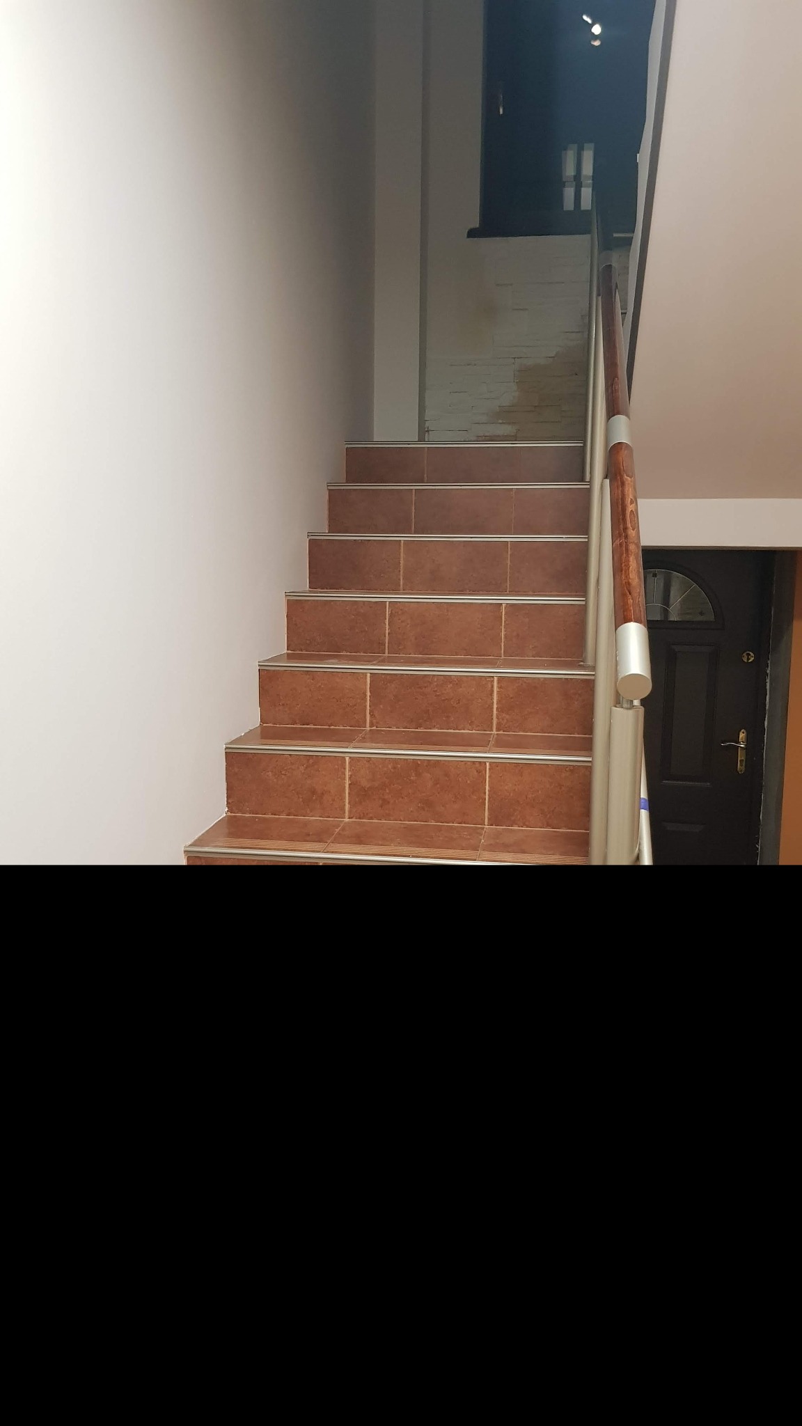 Schody wewnętrzne wyłożone brązowymi płytkami z metalowymi listwami, z drewnianą poręczą i srebrnymi wspornikami, prowadzące do ciemnego pomieszczenia z oknem i pomalowaną na biało ceglaną ścianą...
