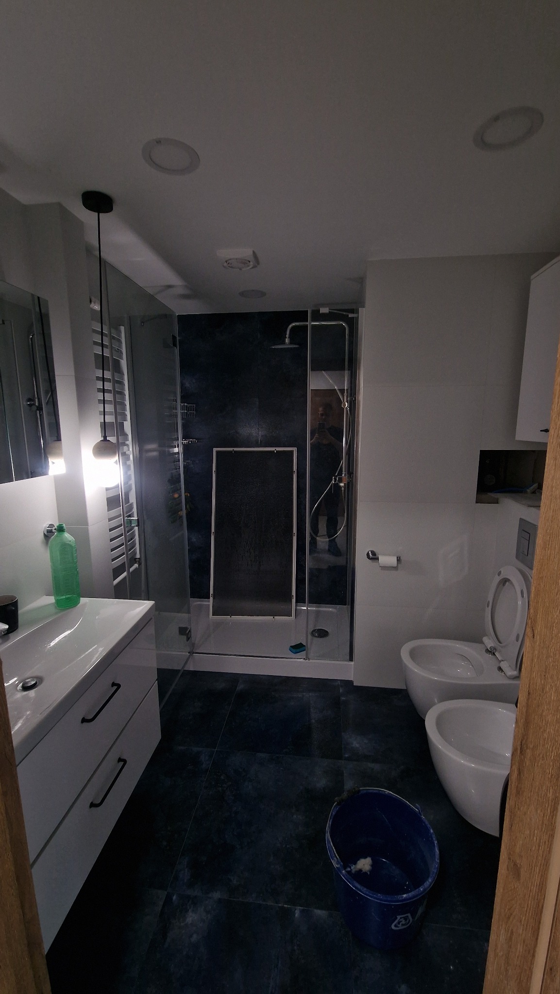 Nowoczesna łazienka z kabiną prysznicową typu walk-in, białą umywalką z szafką i toaletą. Na podłodze ciemne płytki. Widoczne akcesoria łazienkowe i wiadro.