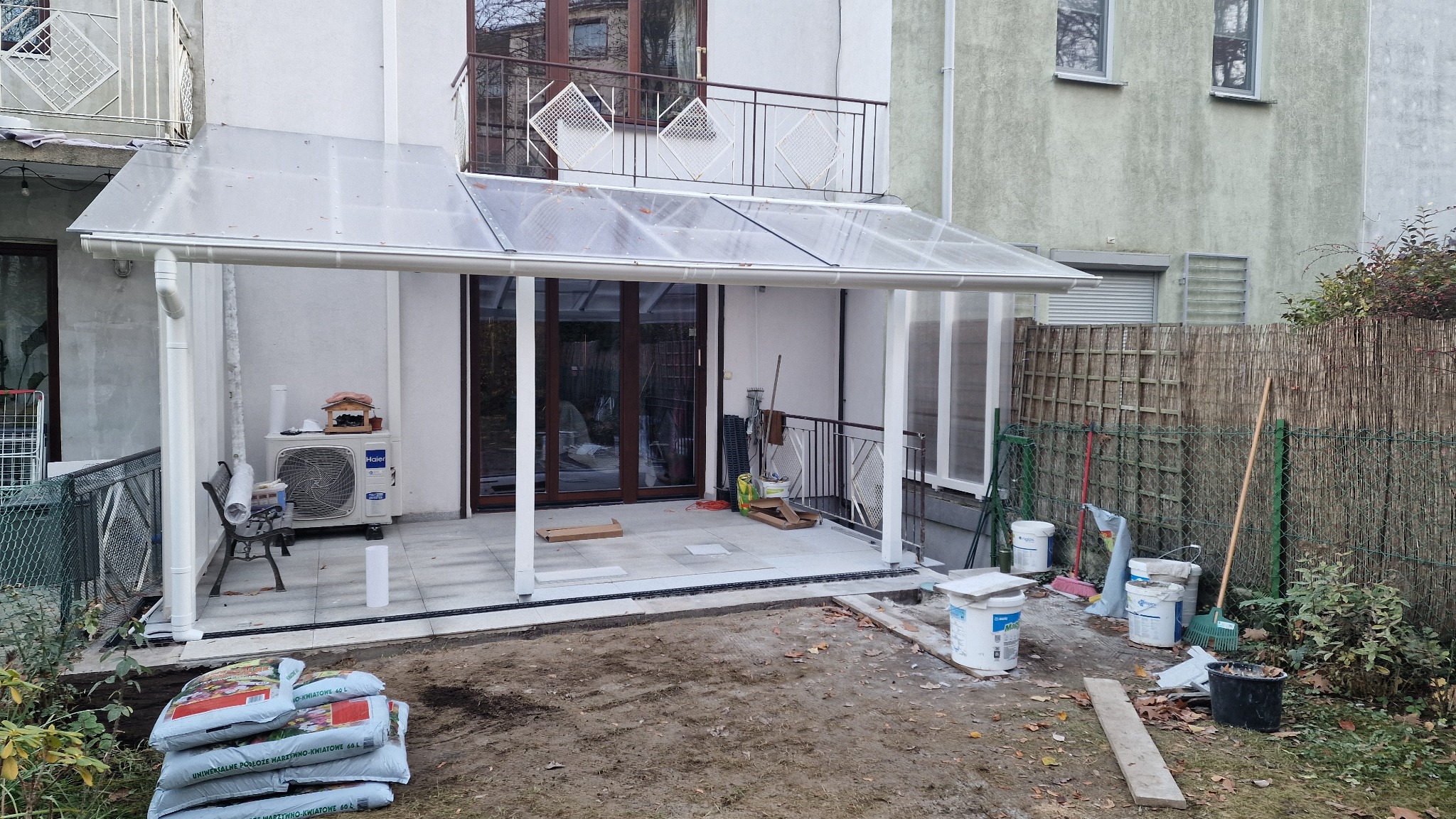 Zadaszony taras z poliwęglanu przy budynku, widoczne worki ziemi i narzędzia. Częściowo zabudowany balkon z widokiem na ogród.