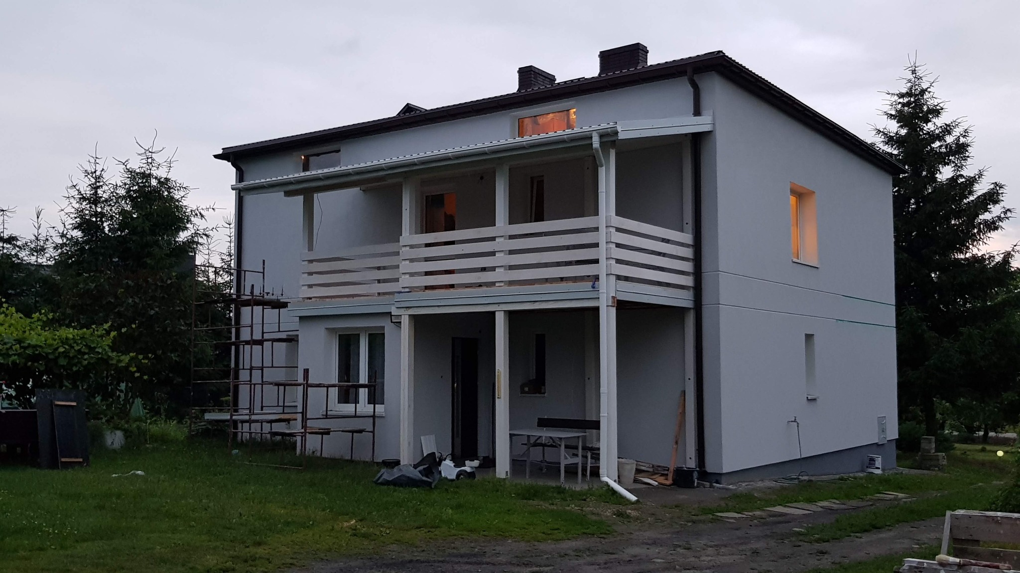 Dwukondygnacyjny dom z białymi balkonami i szarymi ścianami, w trakcie prac wykończeniowych. Widoczne rusztowania i elementy wyposażenia ogrodu.