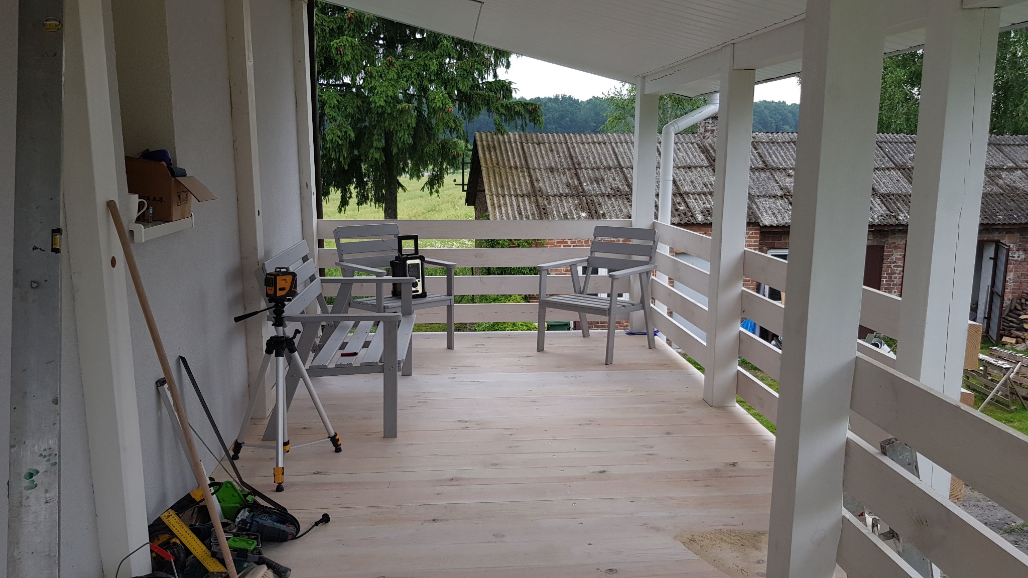 Jasny, odnowiony balkon z drewnianą podłogą i szarymi meblami ogrodowymi. Widoczny laser krzyżowy na statywie i narzędzia, sugerujące prace wykończeniowe.