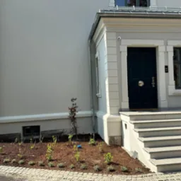 Elewacja domu w jasnych odcieniach szarości z ciemnymi drzwiami wejściowymi i schodami, świeżo założona rabata z korą i młodymi roślinami, widoczny fragment chodnika z kostki brukowej.