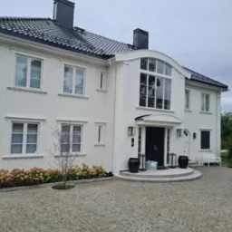 Elegancki, dwukondygnacyjny dom z białymi panelami, czarnym dachem i czarnymi akcentami, takimi jak komin i donice, z brukowanym podjazdem i rabatą z hortensjami.