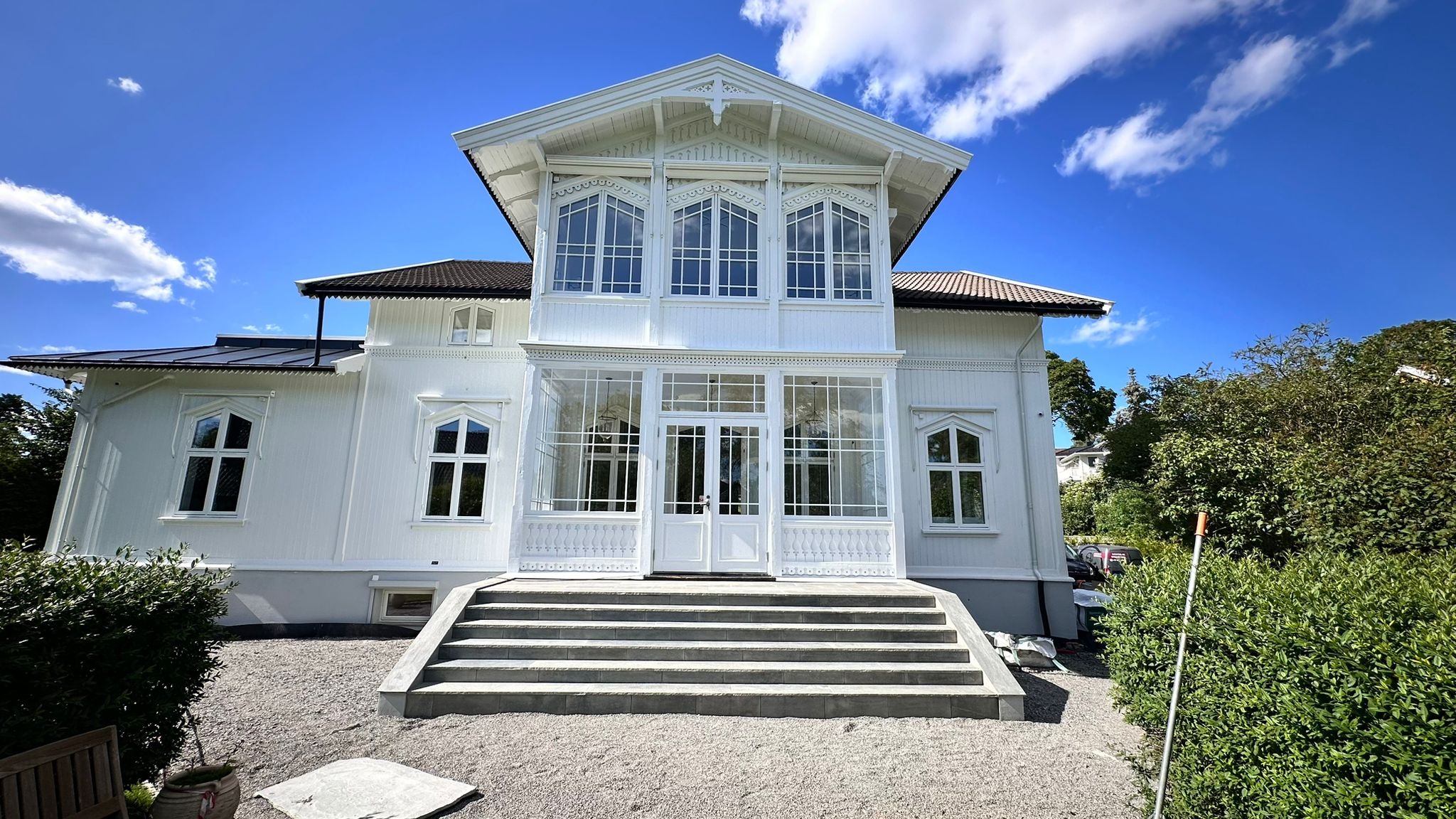 Elegancki, biały dom z werandą i dużymi oknami, widok z przodu na schody wejściowe i żwirowe podłoże, błękitne niebo z chmurami w tle.