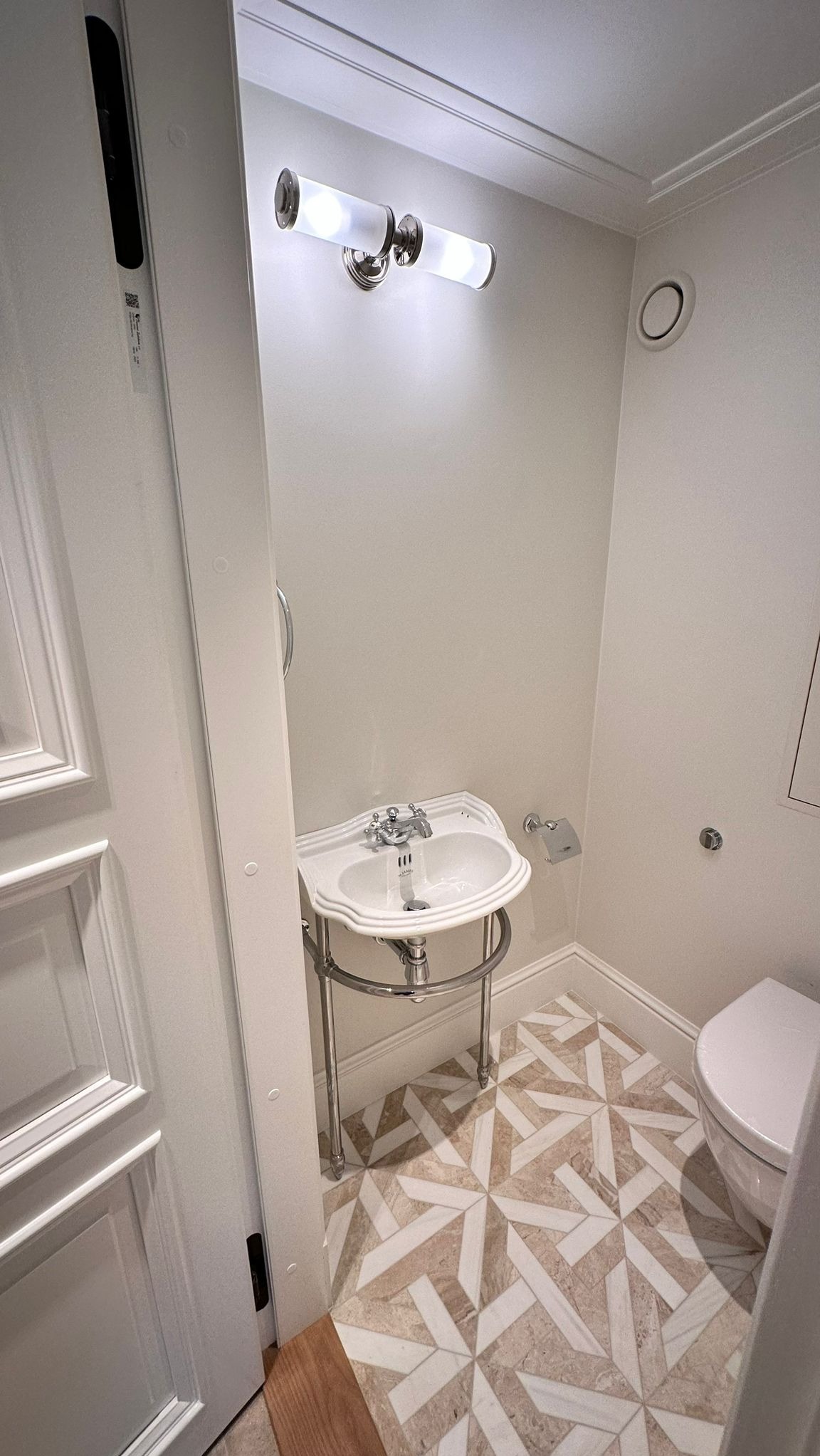 Biała umywalka na chromowanym stelażu z ozdobną armaturą, dekoracyjna mozaika podłogowa w beżowo-białej kolorystyce, oświetlenie punktowe w łazience.