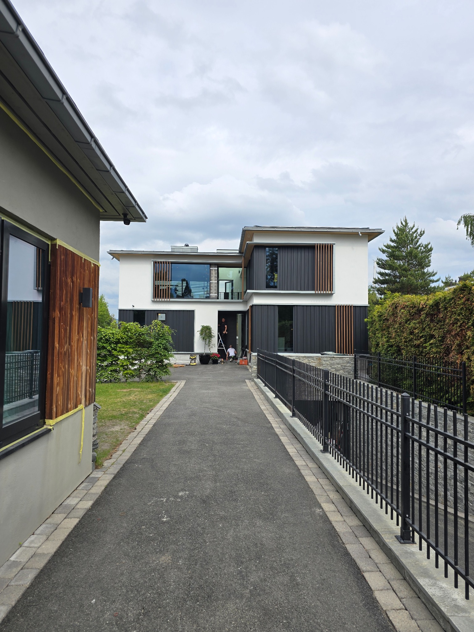 Nowoczesny dom z płaskim dachem i elementami drewnianymi, widoczni pracownicy na drabinie, przygotowania do prac elewacyjnych, ścieżka prowadząca do wejścia.