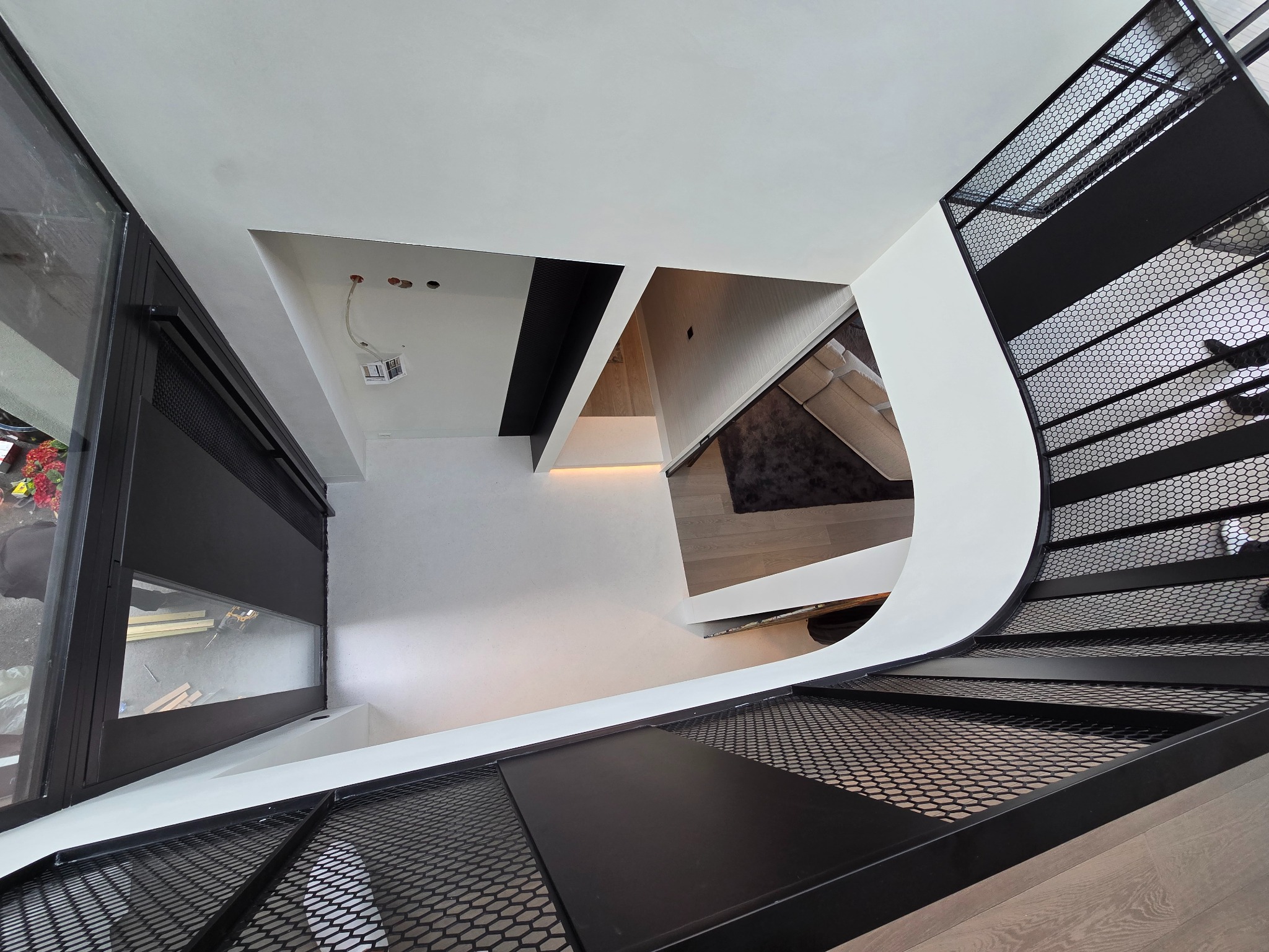 Wnętrze domu z nowoczesnymi schodami o metalowej konstrukcji z siatki, widok z góry na salon z kominkiem i częściowo widocznym umeblowaniem.